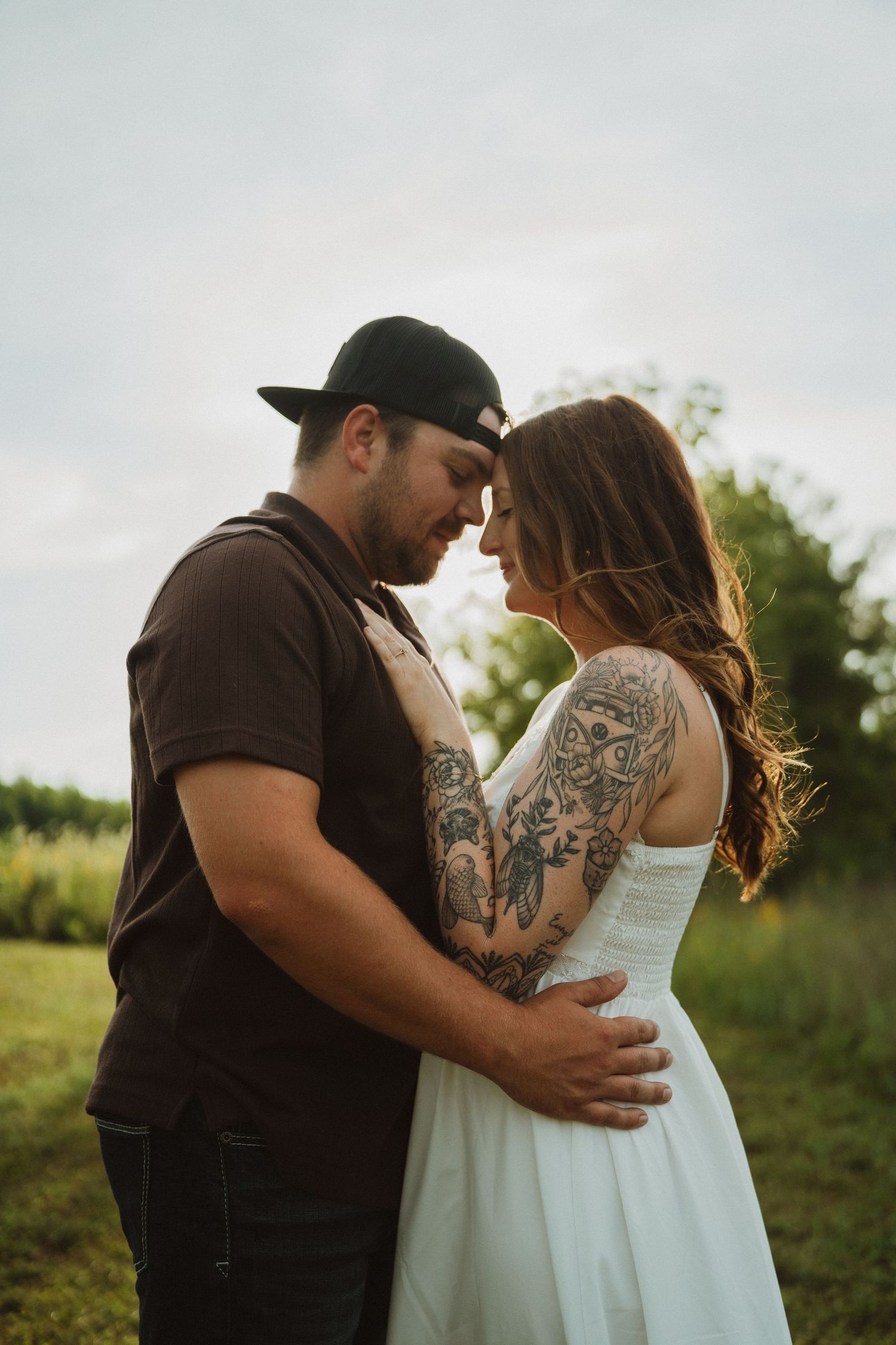 Couple embracing in a field, woman in white dress with tattoos, man in dark shirt and cap, romantic outdoor portrait.