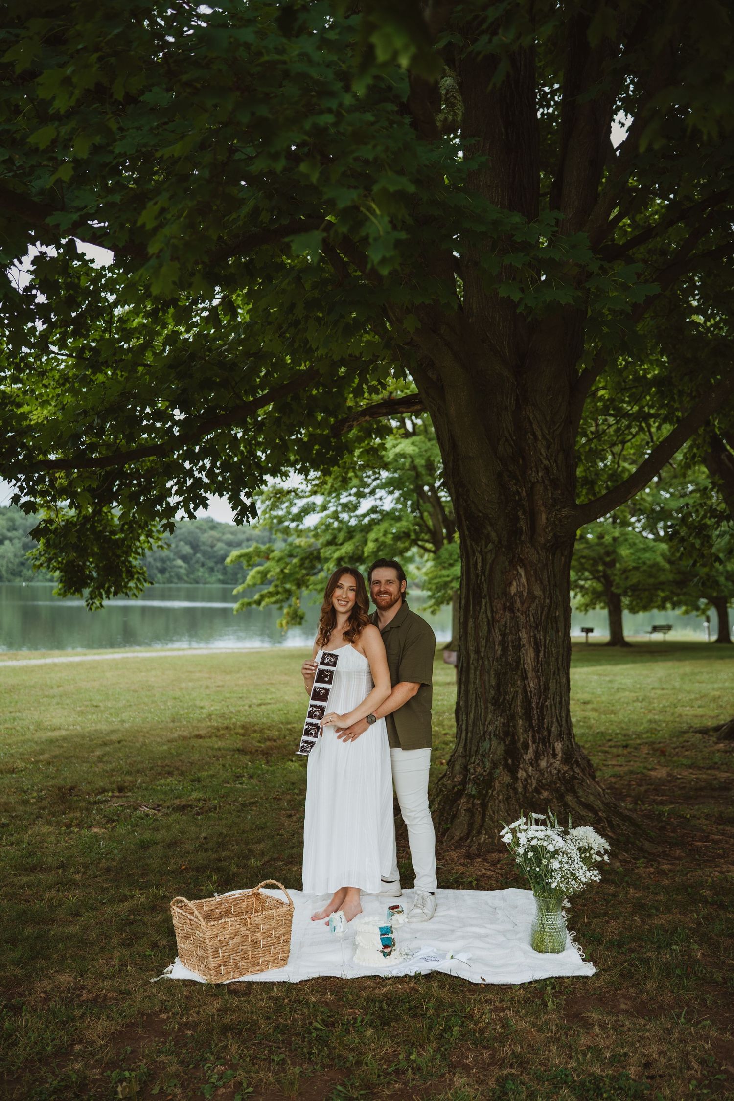 Couple standing under a large tree holding ultrasound photos for a pregnancy announcement at a lakeside park picnic.