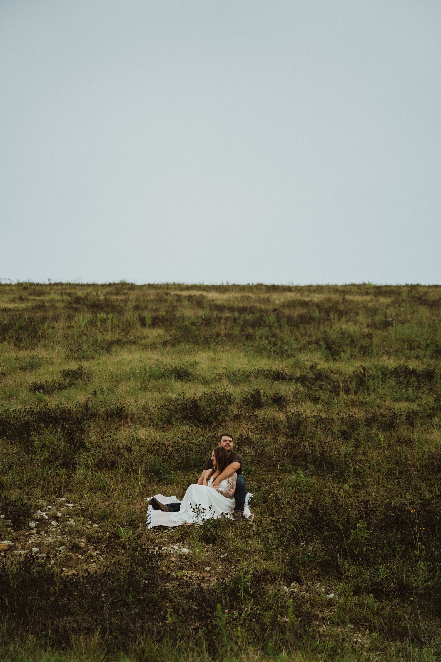 Couple sitting together in a lush green meadow, woman in white dress, man looking up at the sky.