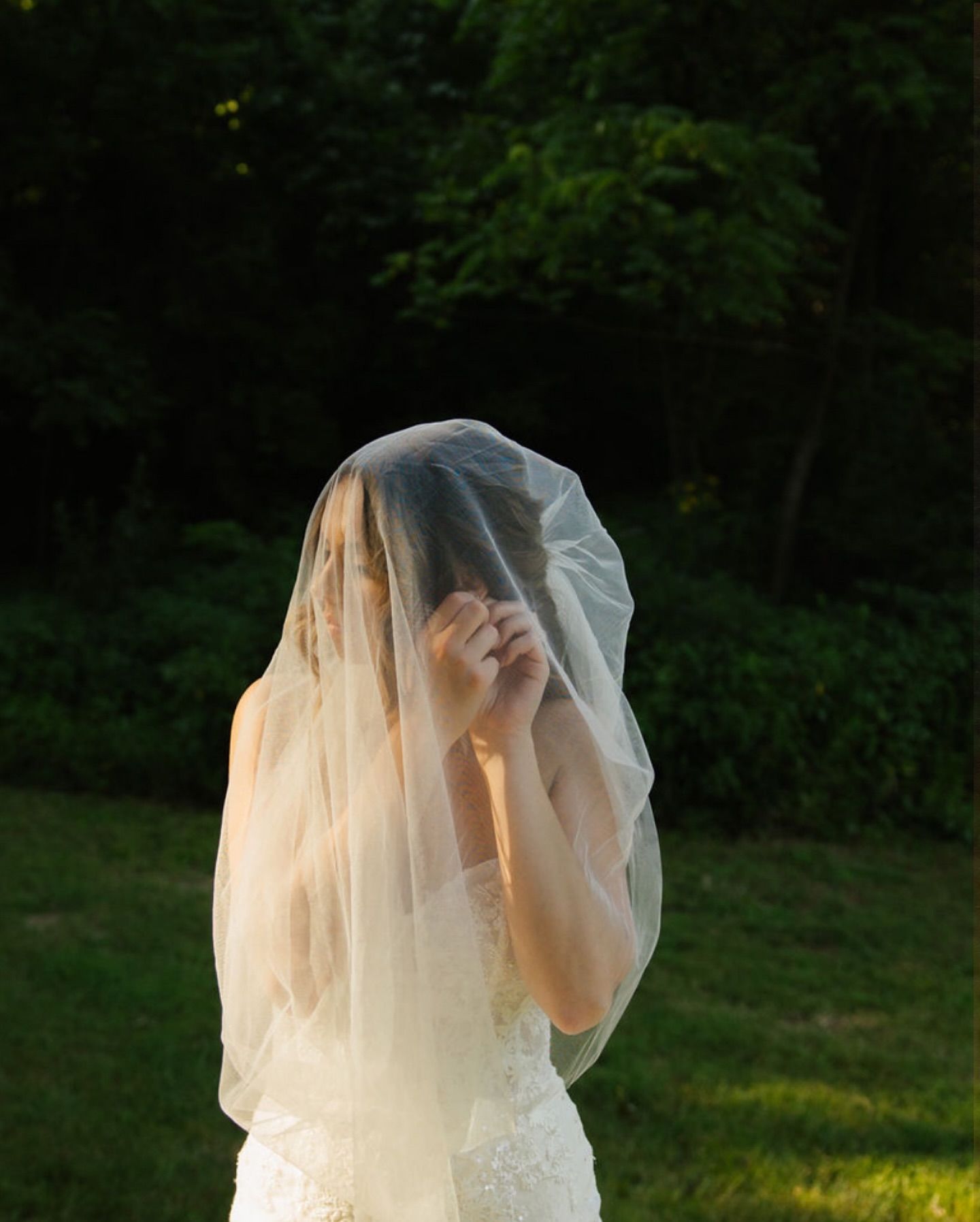 A bride in a flowing white veil stands against a lush green forest backdrop during golden hour.