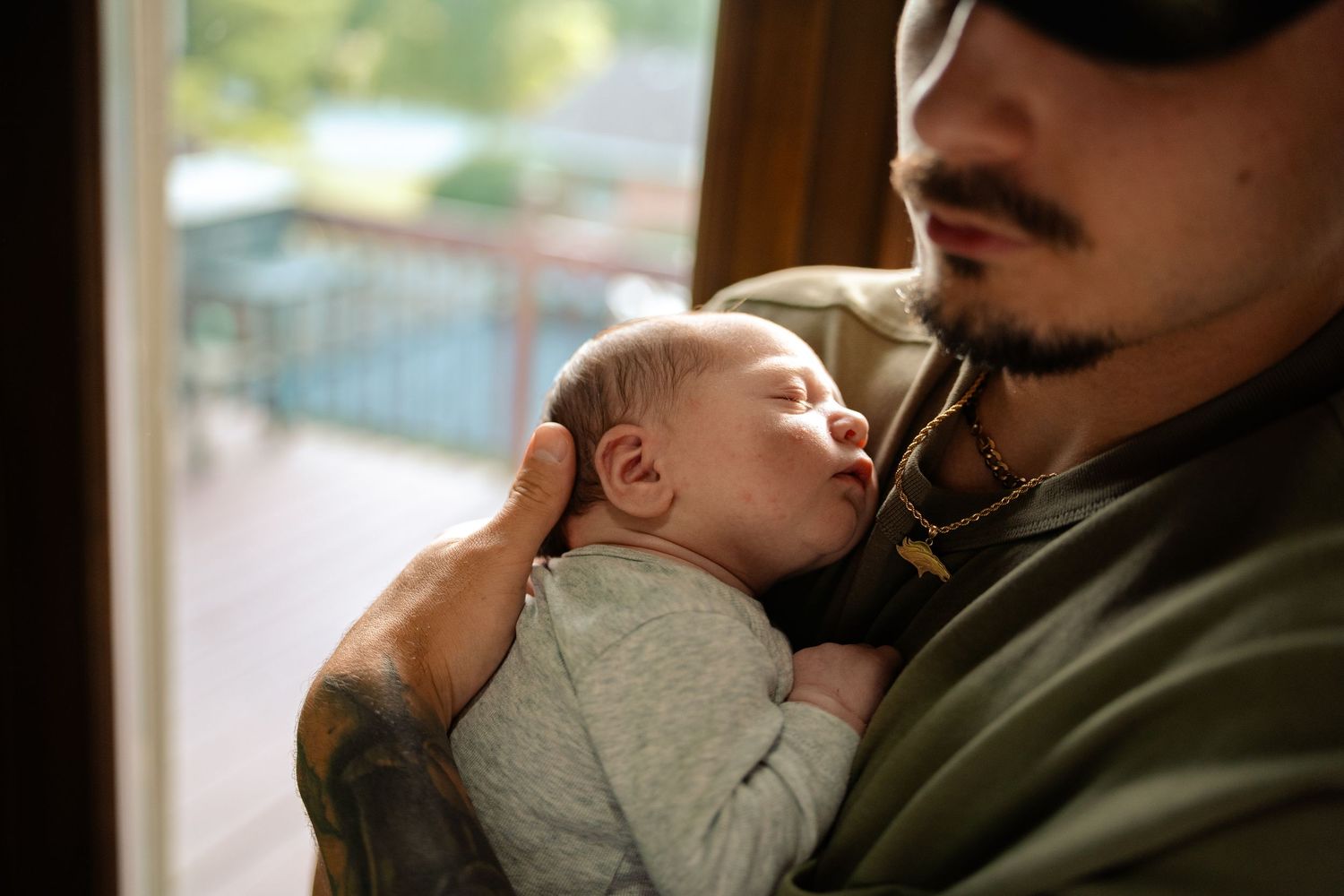 A tattooed father tenderly holds his sleeping newborn baby close to his chest near a window.