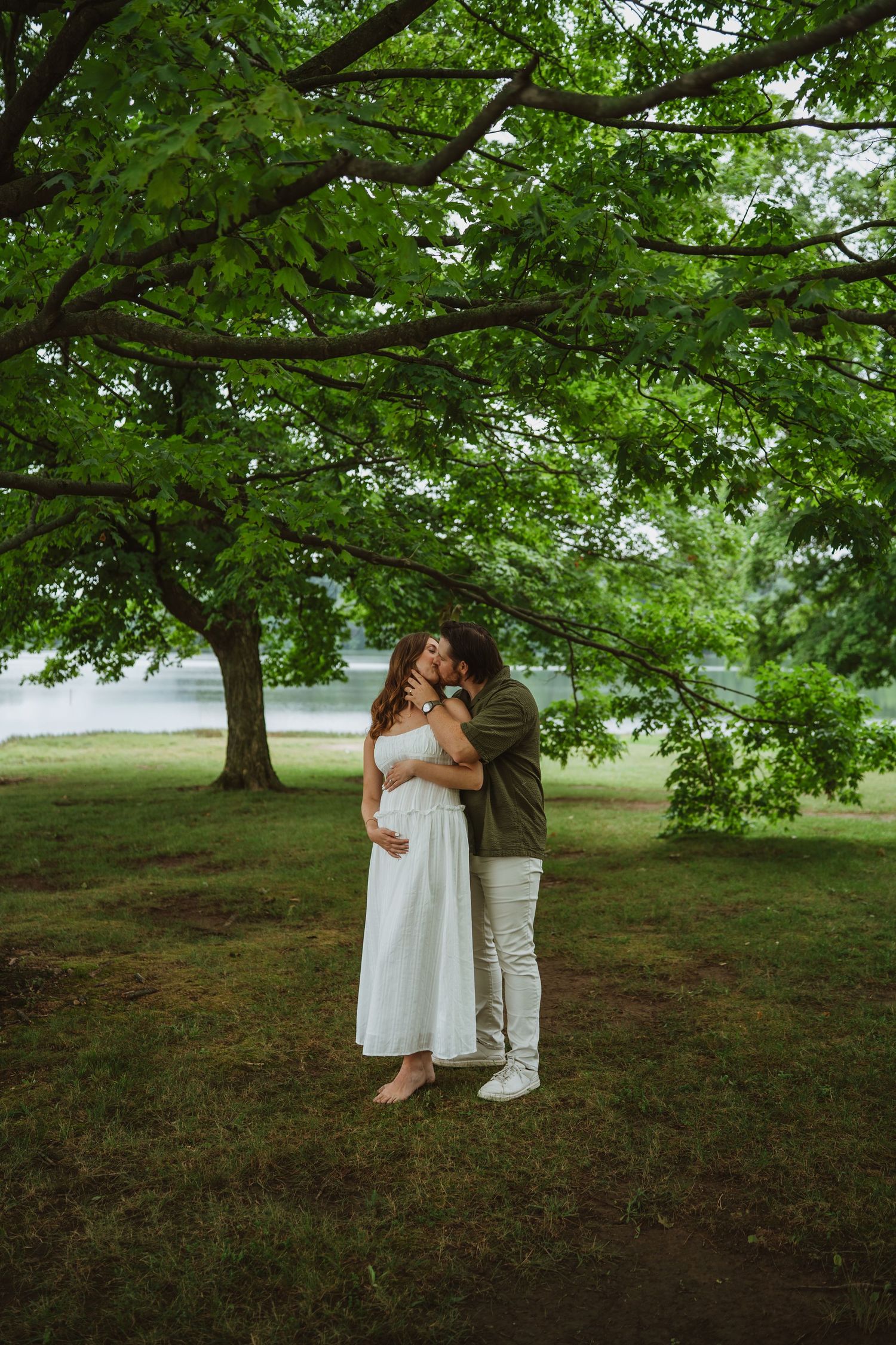 Couple embracing tenderly under lush green trees near a lake, woman in white dress with visible baby bump.