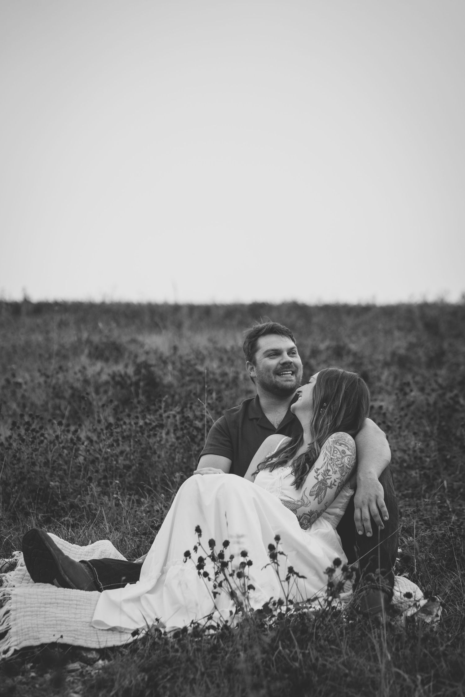 Black and white photo of a couple sitting in a field, woman kissing mans cheek as he smiles, romantic outdoor portrait.