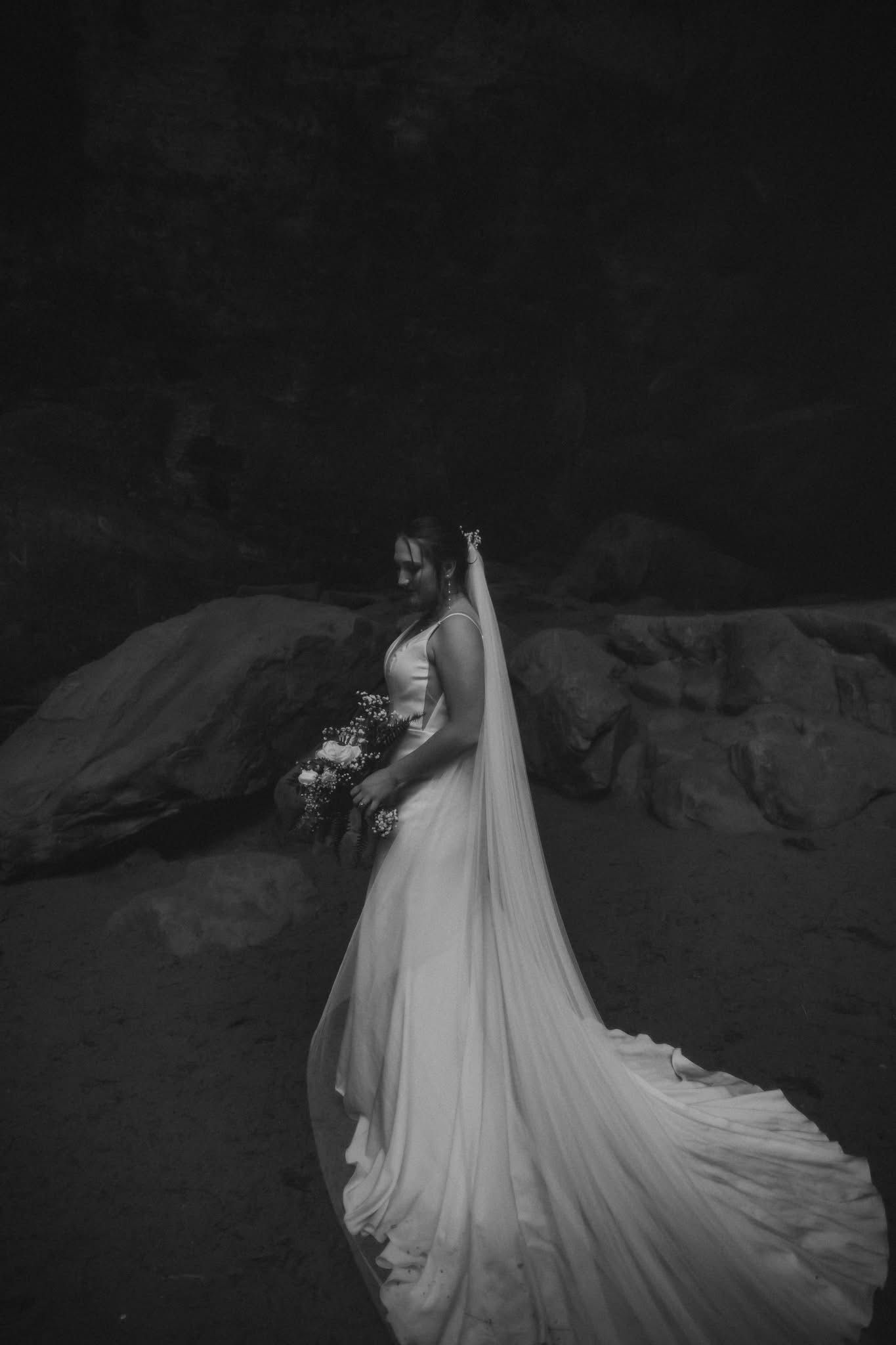 Black and white bridal portrait of a bride in a flowing gown and veil holding a bouquet outdoors.