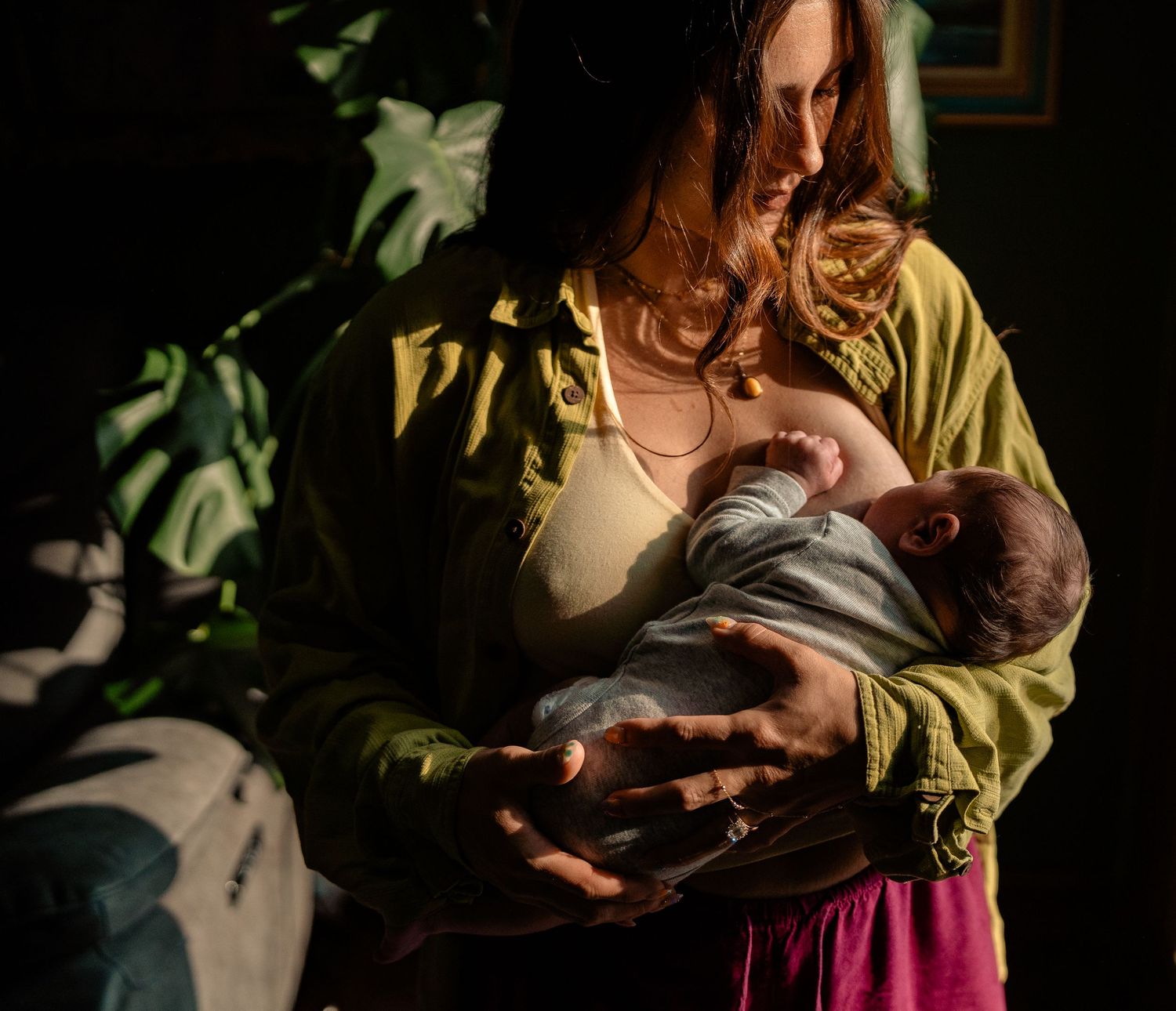 Mother in olive green top breastfeeds newborn baby in dramatic moody natural light at home.