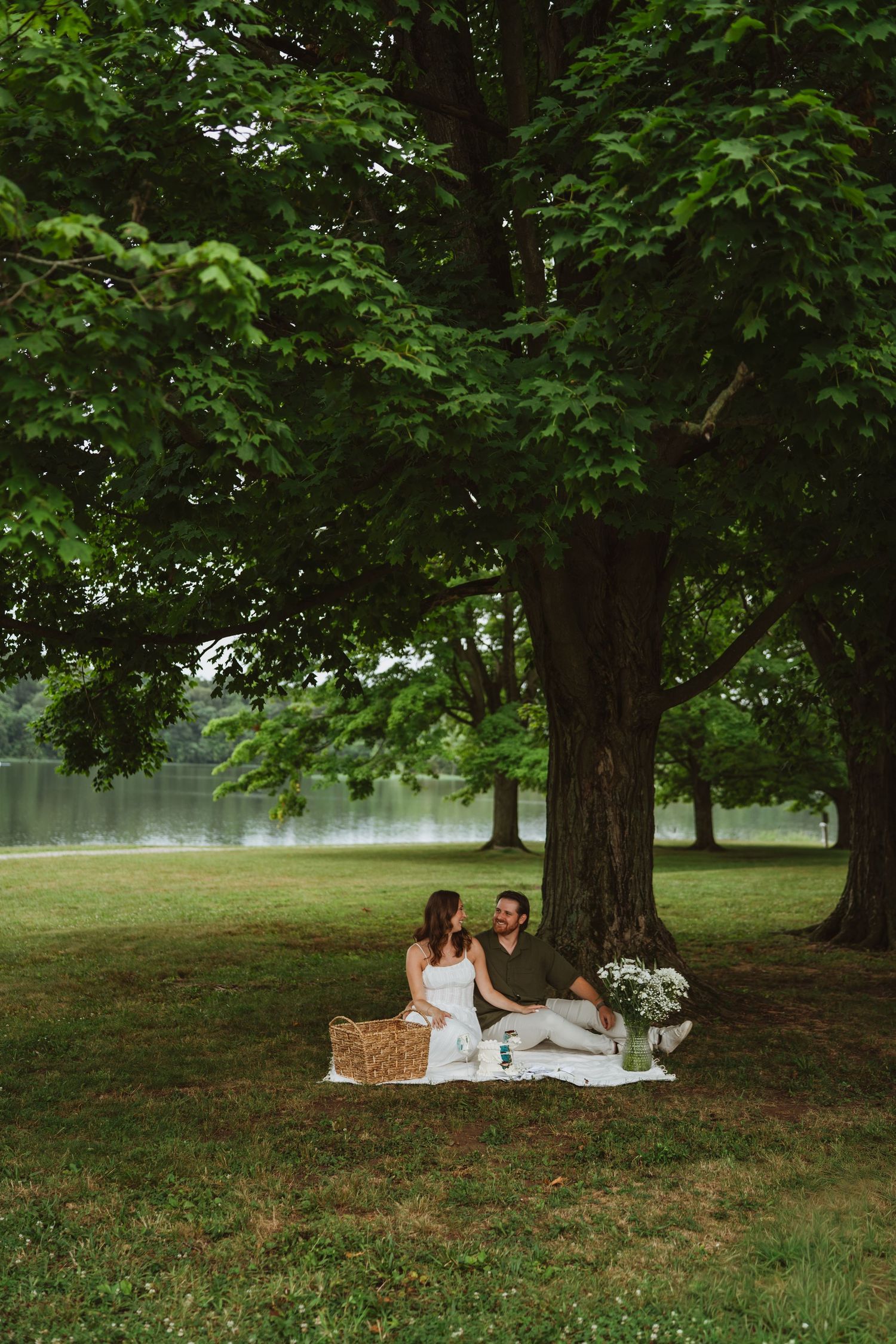 Couple enjoying a romantic picnic on a blanket under a large tree by a serene lake in a lush green park.
