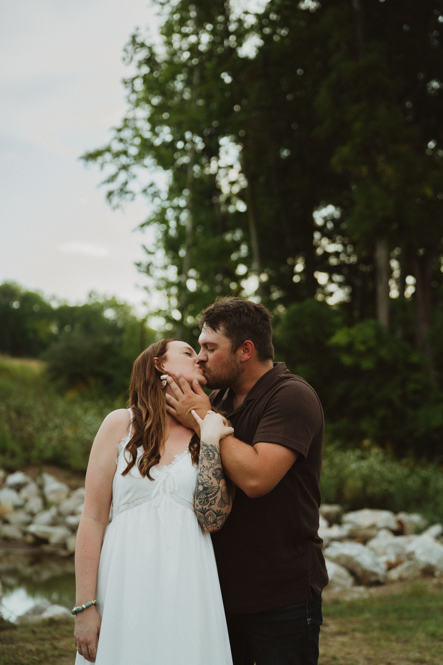 A couple shares a romantic kiss outdoors, man with tattooed arm in black shirt, woman in white dress, trees behind.