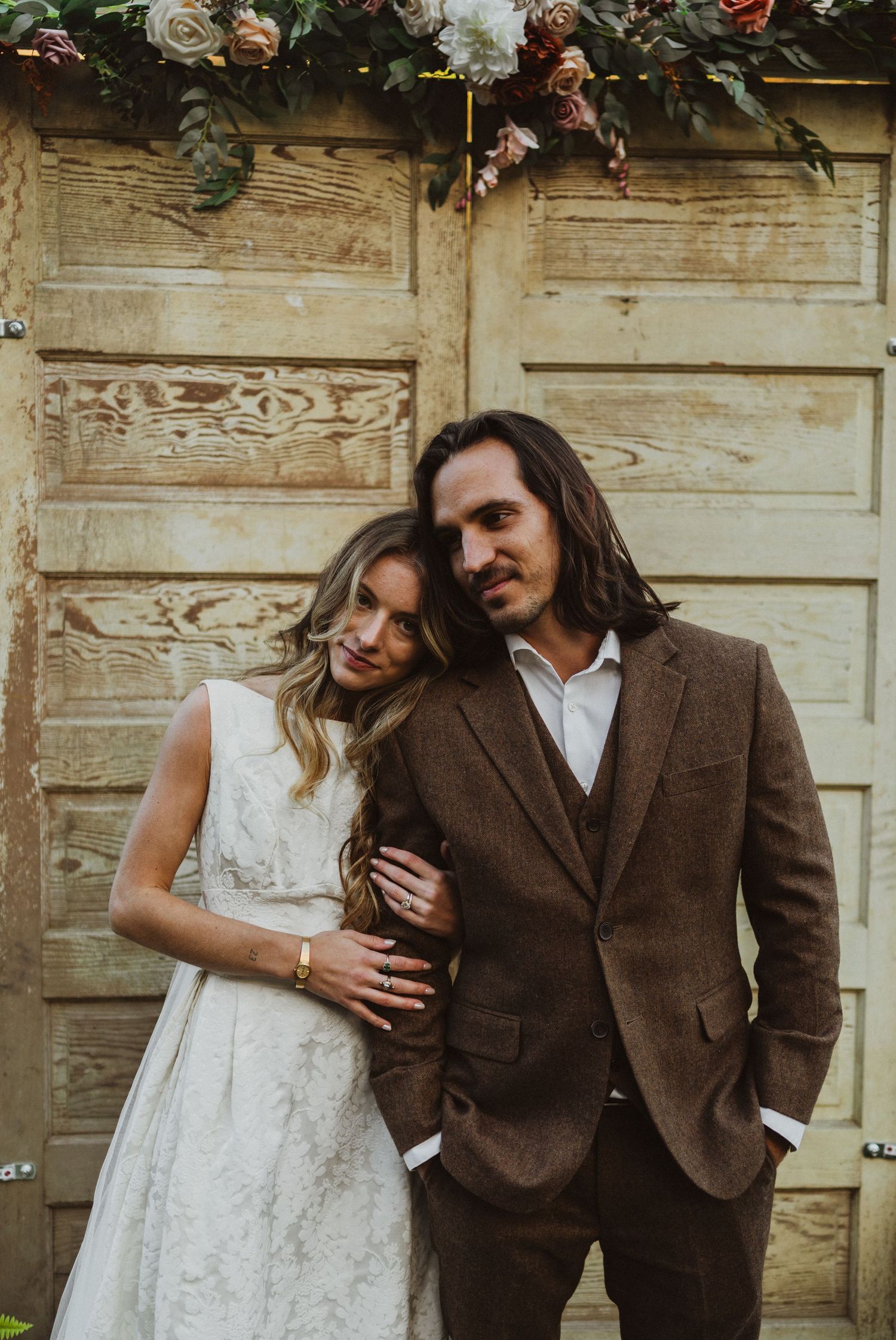 Romantic couple in vintage attire poses against weathered wooden doors in sepia tones.