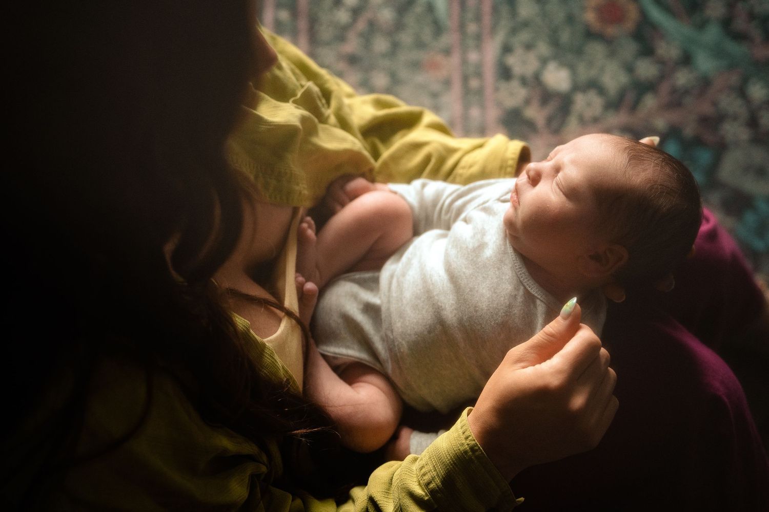 A mother tenderly holds her sleeping newborn baby wrapped in white cloth in a dimly lit room.