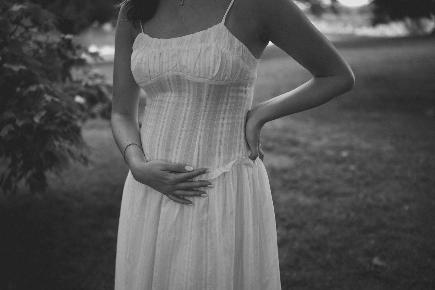 Black and white photo of pregnant woman in white dress cradling baby bump outdoors.