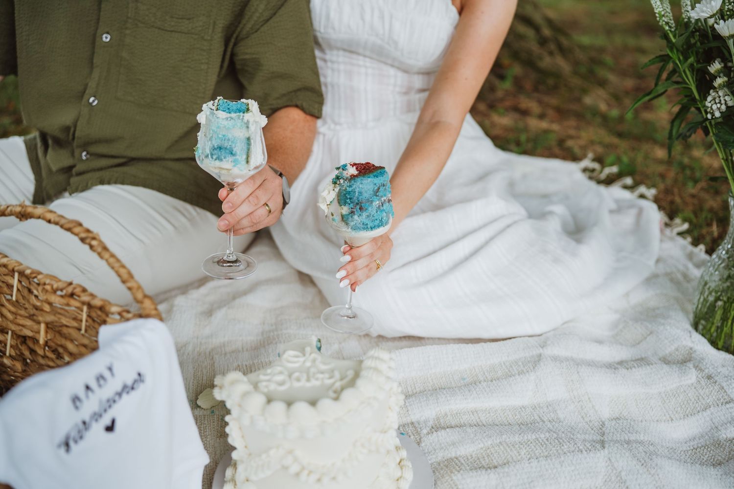Couple holding blue-painted wine glasses at a picnic with a white cake and wicker basket on a blanket.