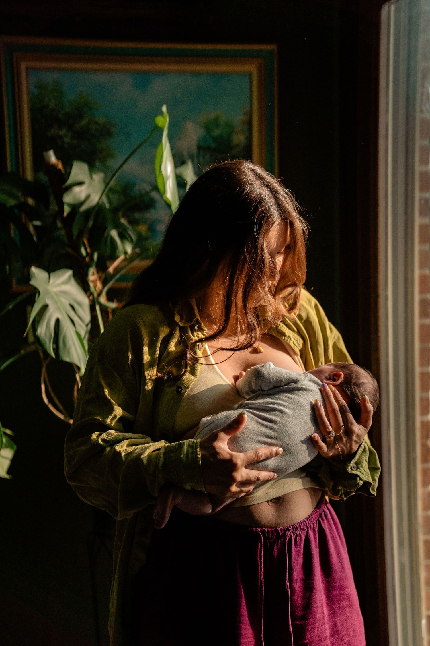 A mother breastfeeding her newborn baby in warm sunlight near a window with lush green plants nearby.