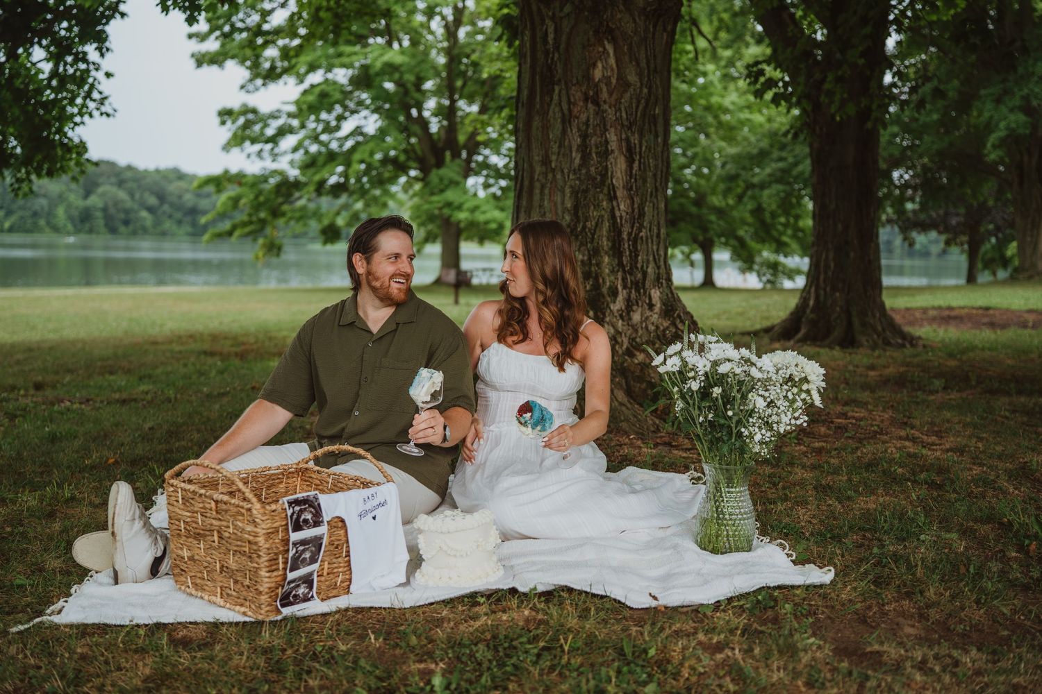 Happy couple enjoying an outdoor picnic near a lake, with a wicker basket and white wildflowers on a blanket.