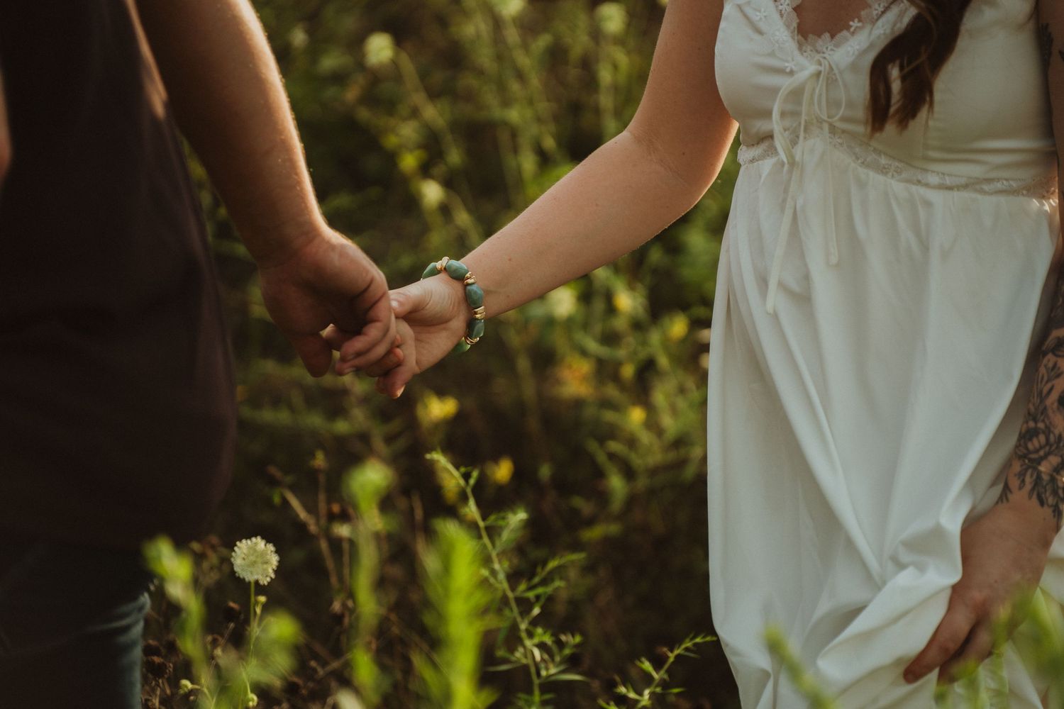 Couple holding hands in a sunlit garden, woman in white dress with bracelet, romantic outdoor engagement photo.