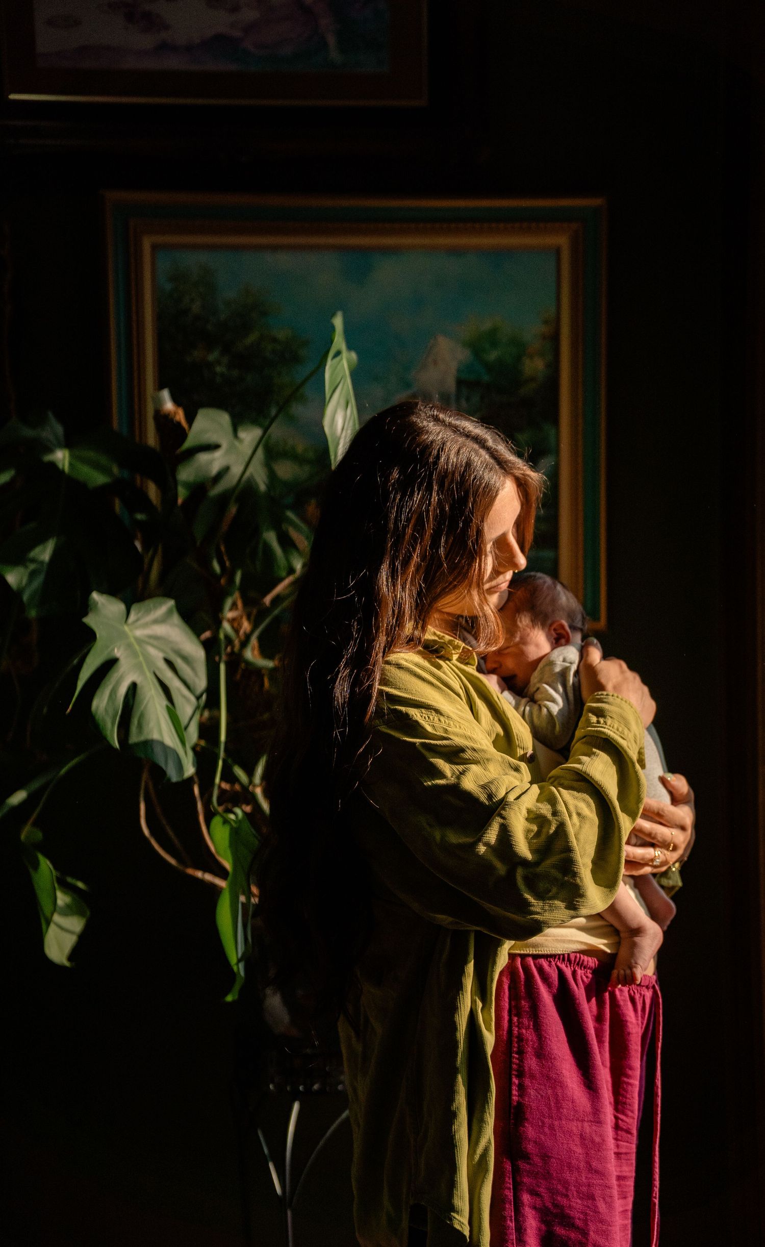 Mother in green jacket holds newborn baby in warm sunlight with monstera plant and painting in background.