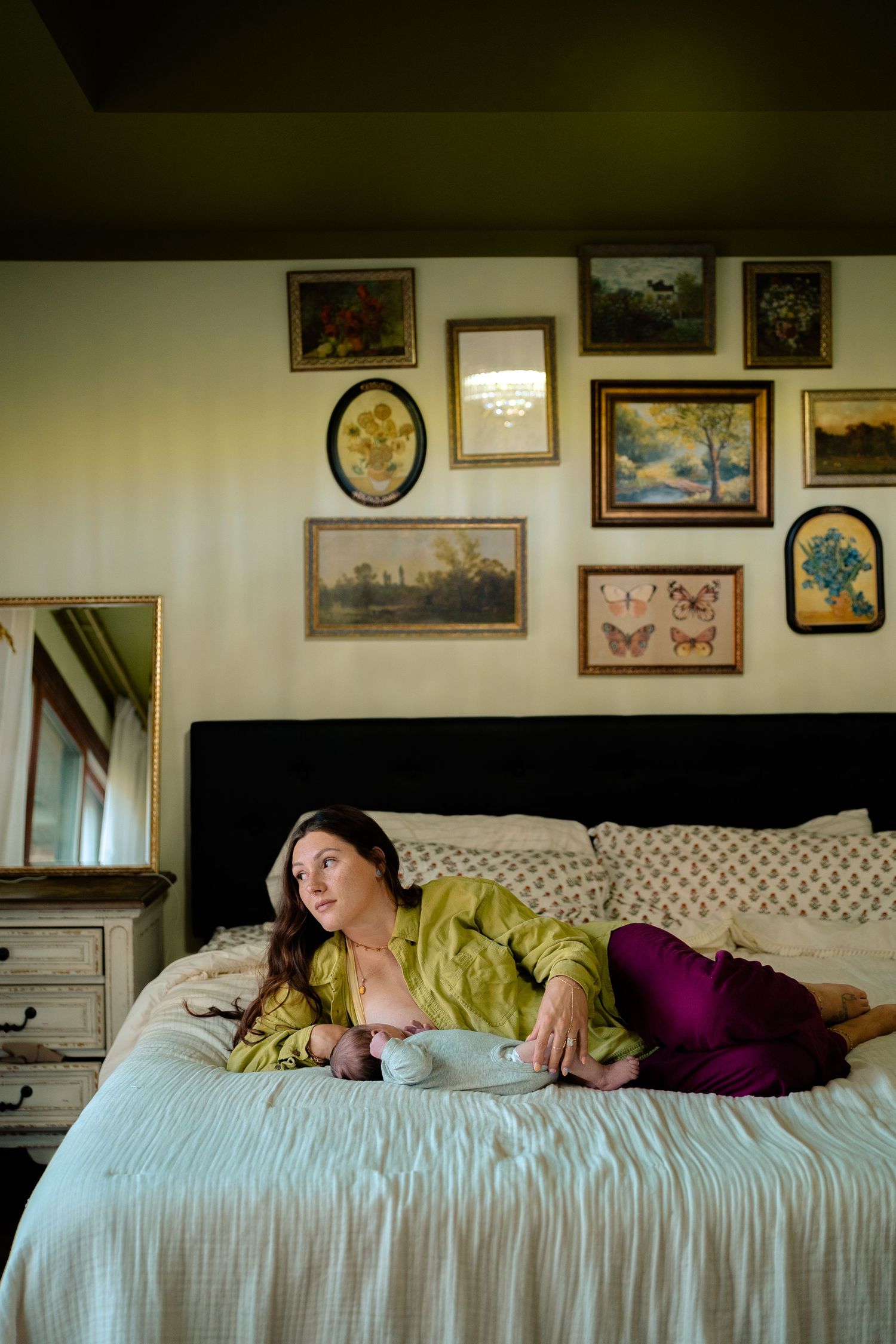 Mother in green silk top reclines on bed with newborn, gallery wall behind her.