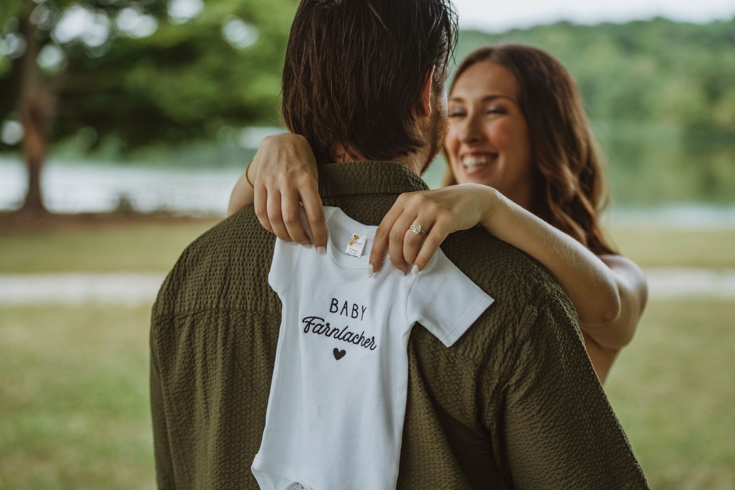 Smiling woman hugging a man while holding a white baby onesie reading Baby Fabricske outdoors.