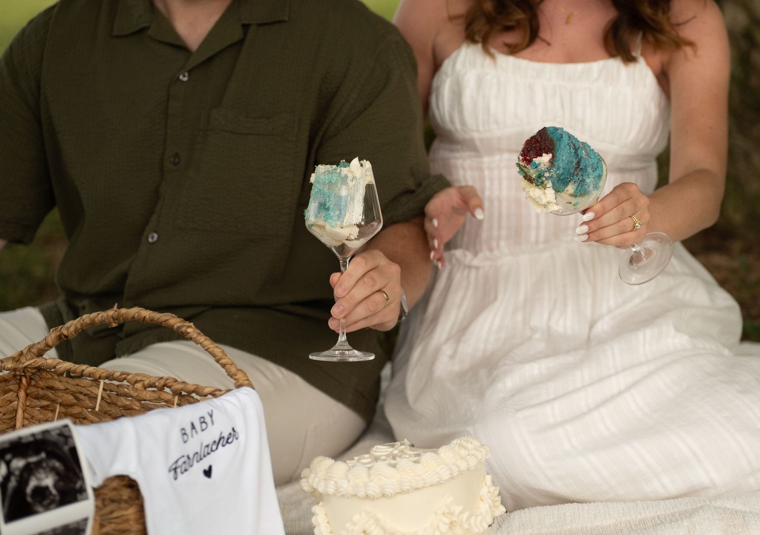 Couple holding wine glasses with ultrasound photo and white cake at a baby announcement picnic.