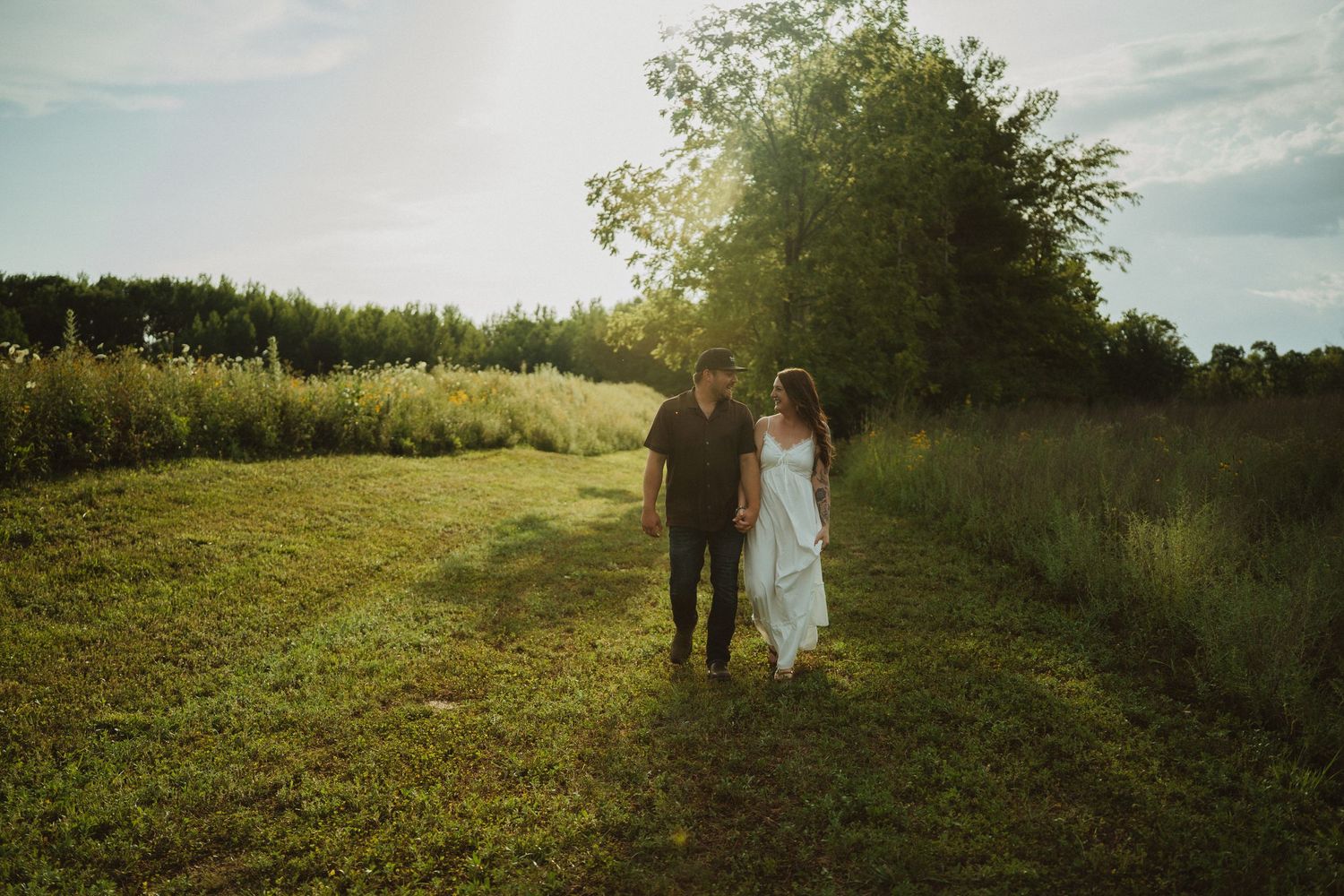 Couple walking hand in hand through a sunlit green meadow at golden hour, woman in white dress, man in dark shirt.