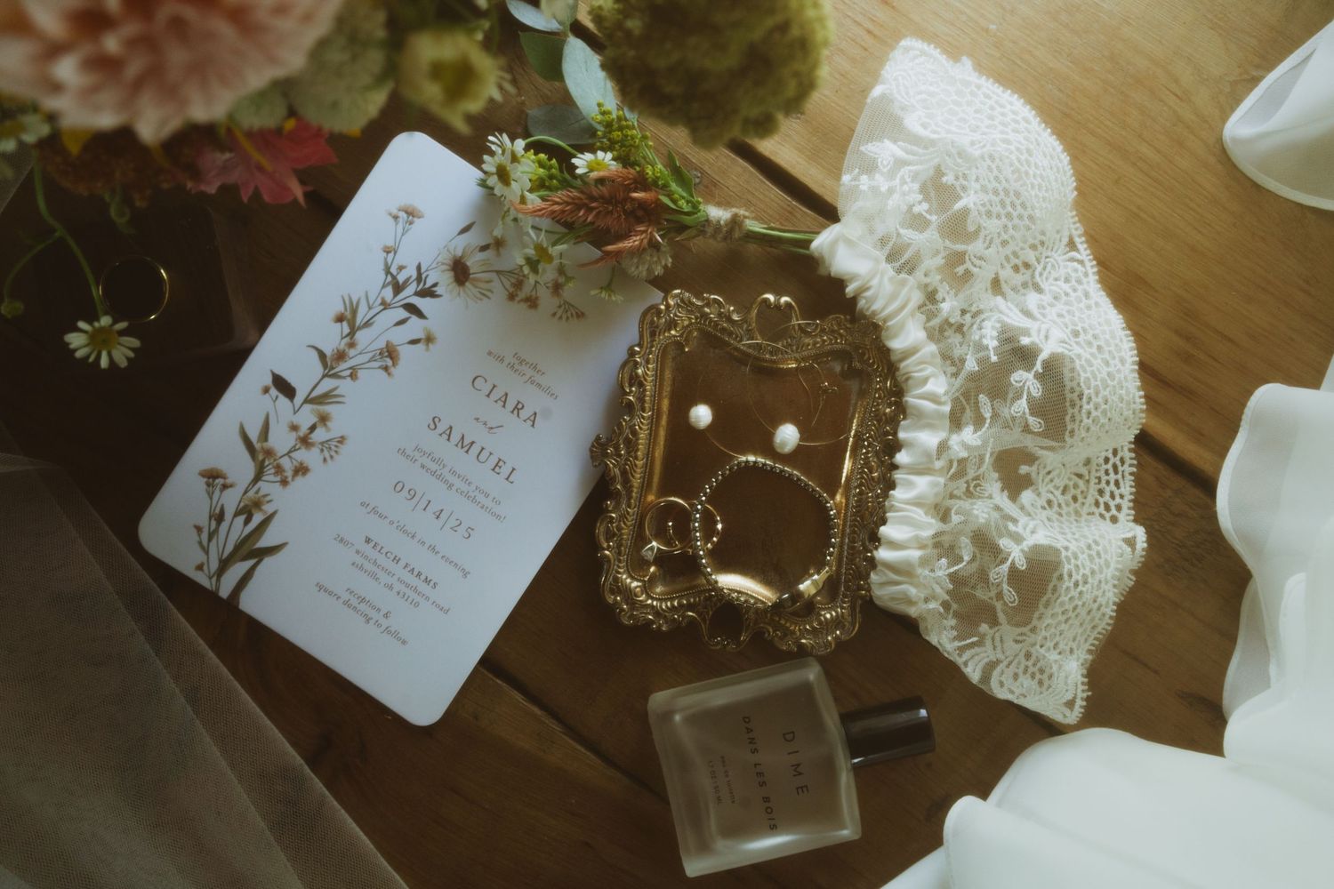 Elegant wedding flat lay with floral invitation, gold ring dish, pearl earrings, perfume bottle, and lace glove on wooden table.