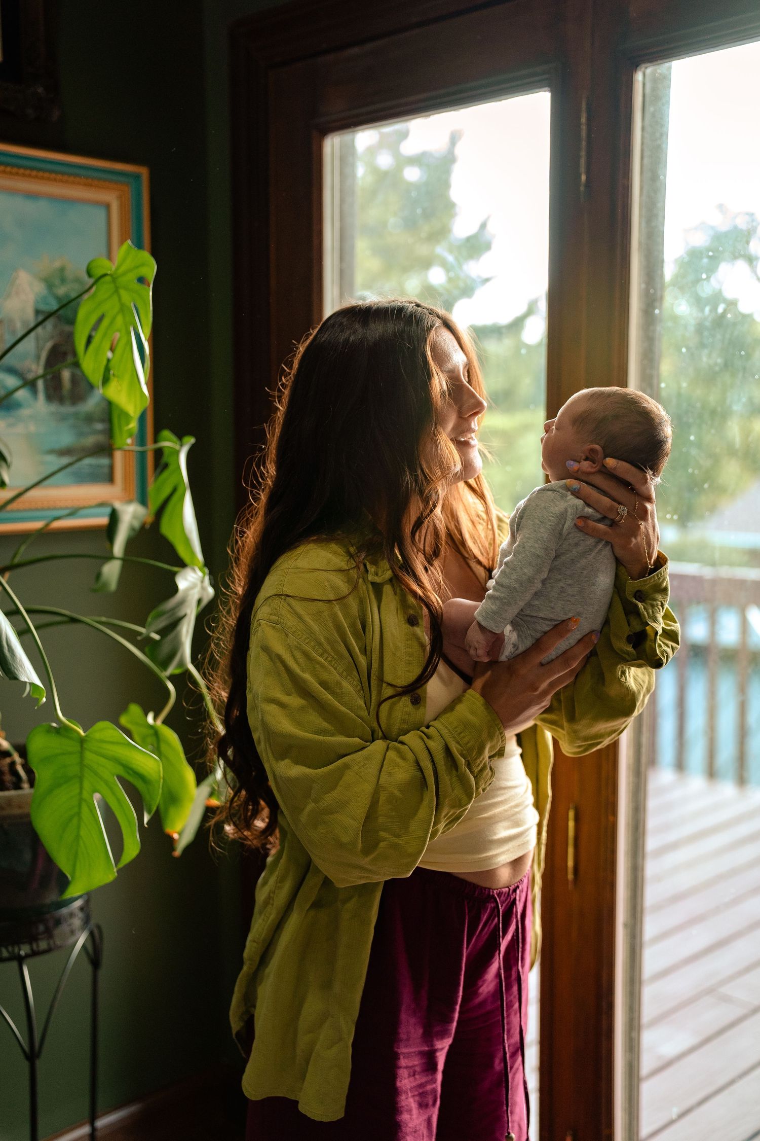 Smiling mother holding newborn baby near sunlit window with monstera plant indoors.