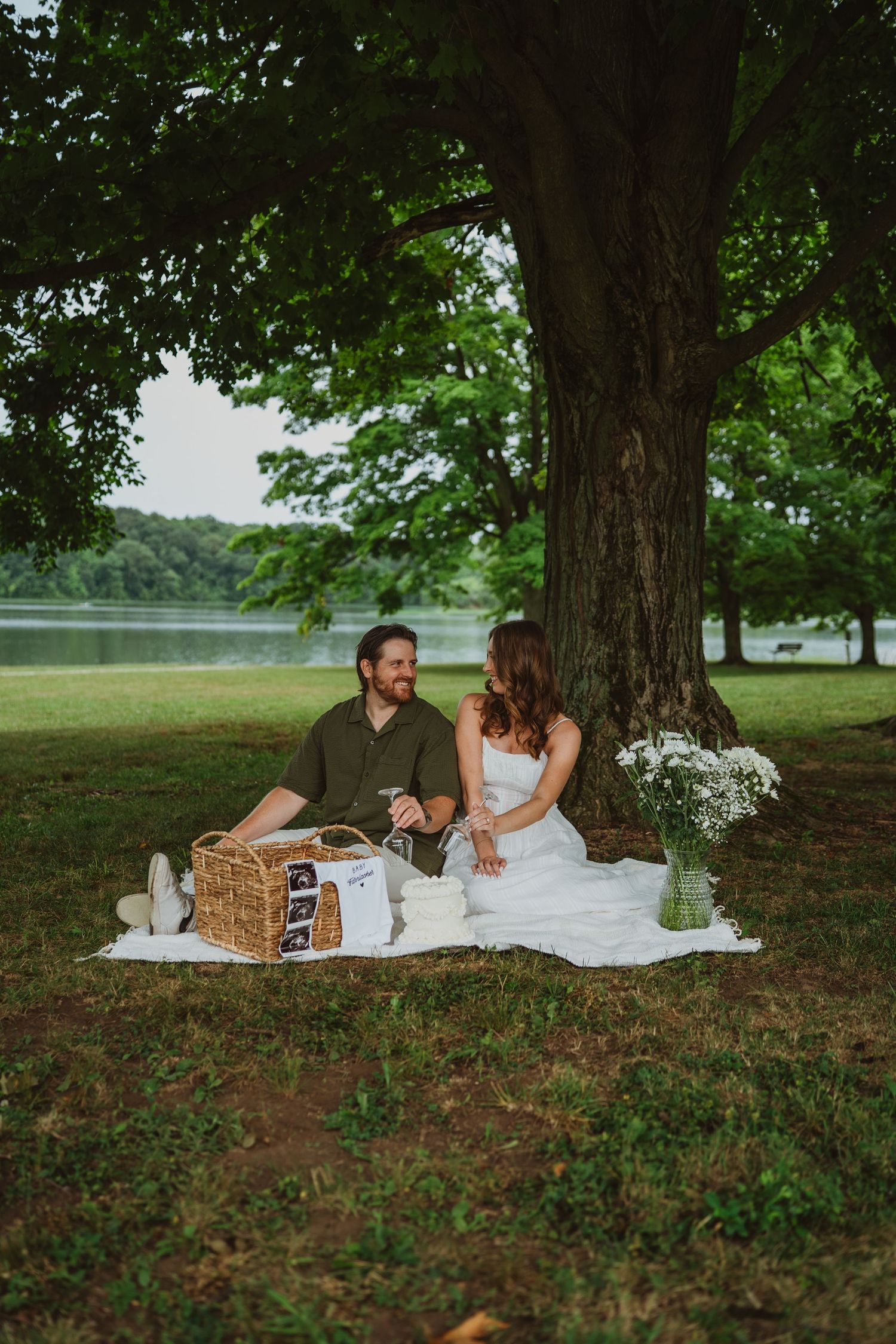 Couple sharing a romantic picnic under a large tree by a lake, sitting on a white blanket with a wicker basket.