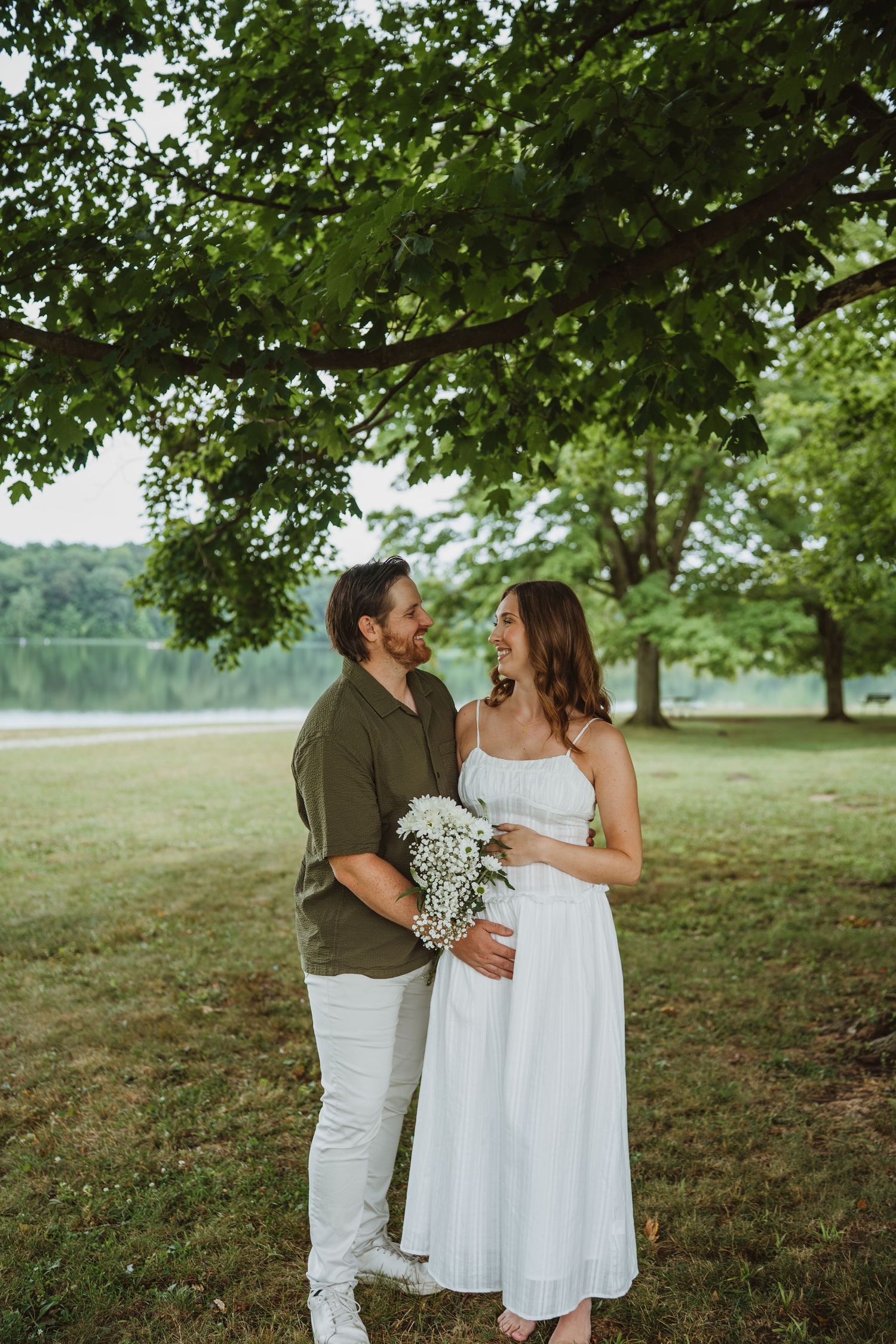 Happy couple standing under tree near lake, man in olive shirt holding white flowers, woman in white dress.