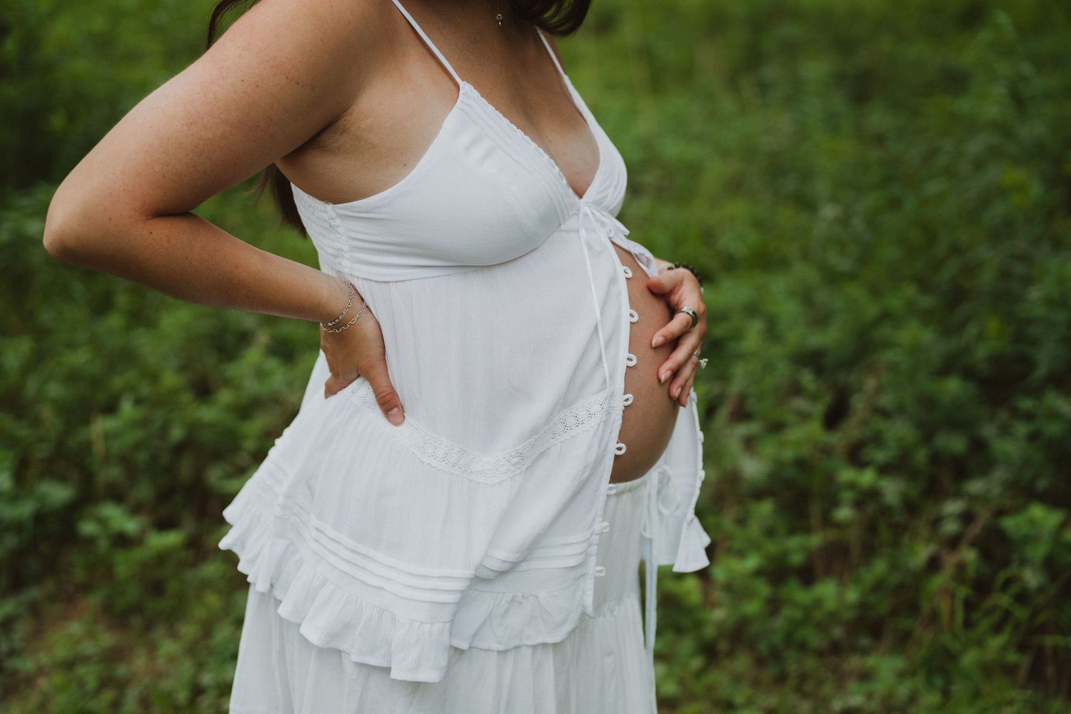 Pregnant woman in white ruffled dress holding baby bump outdoors in green nature setting.