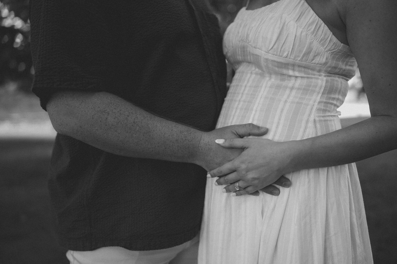 Black and white photo of couple holding hands over pregnant belly outdoors.