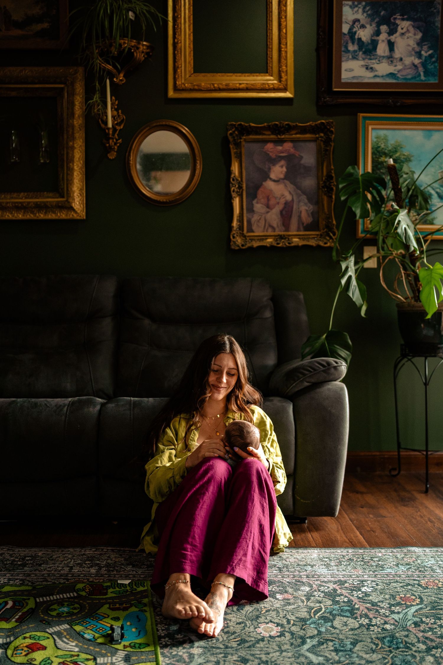 Woman in green top and magenta pants sits on floor holding newborn baby in bohemian living room.