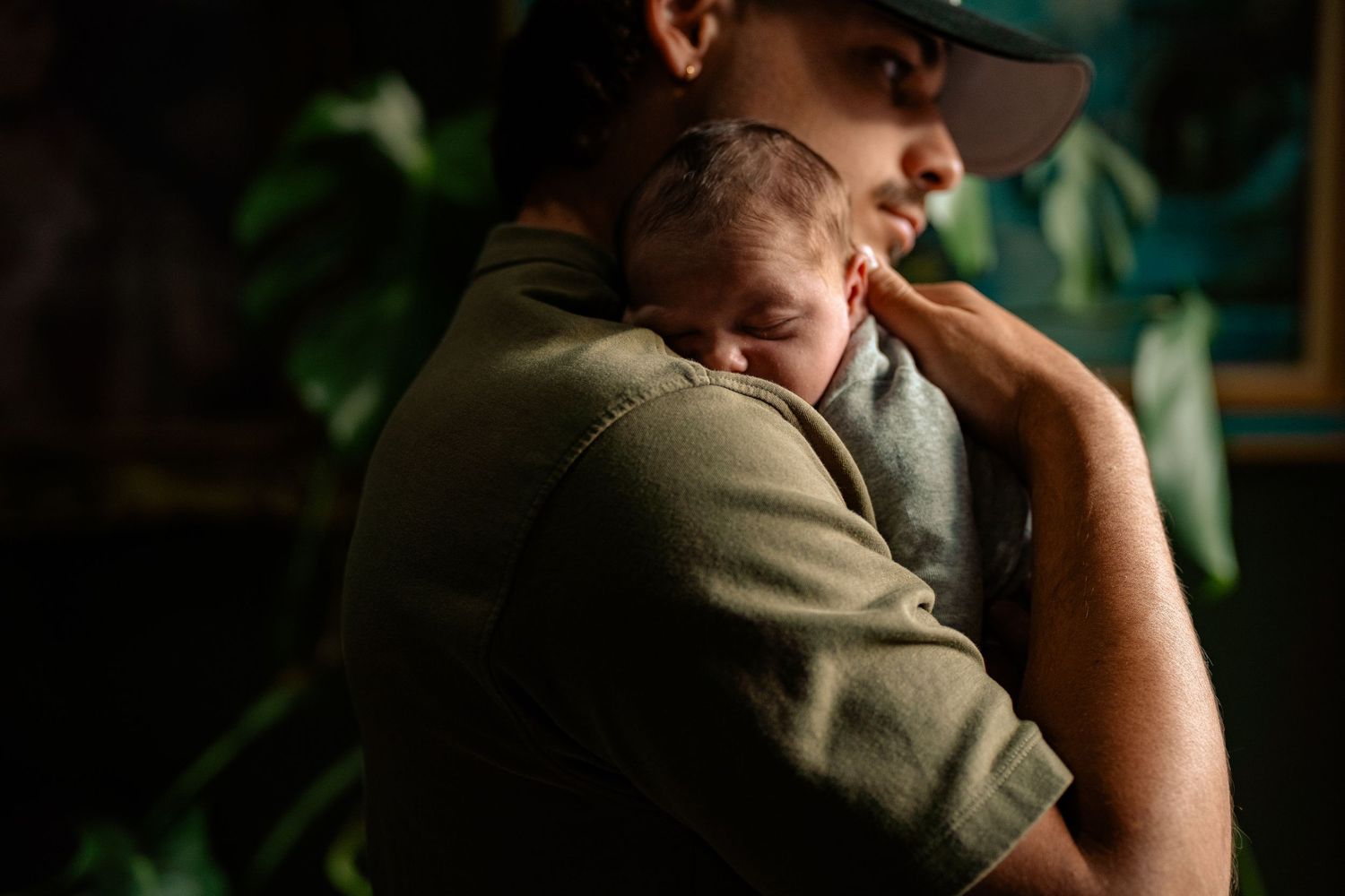 Father in green shirt and cap tenderly holding sleeping newborn baby against his chest.