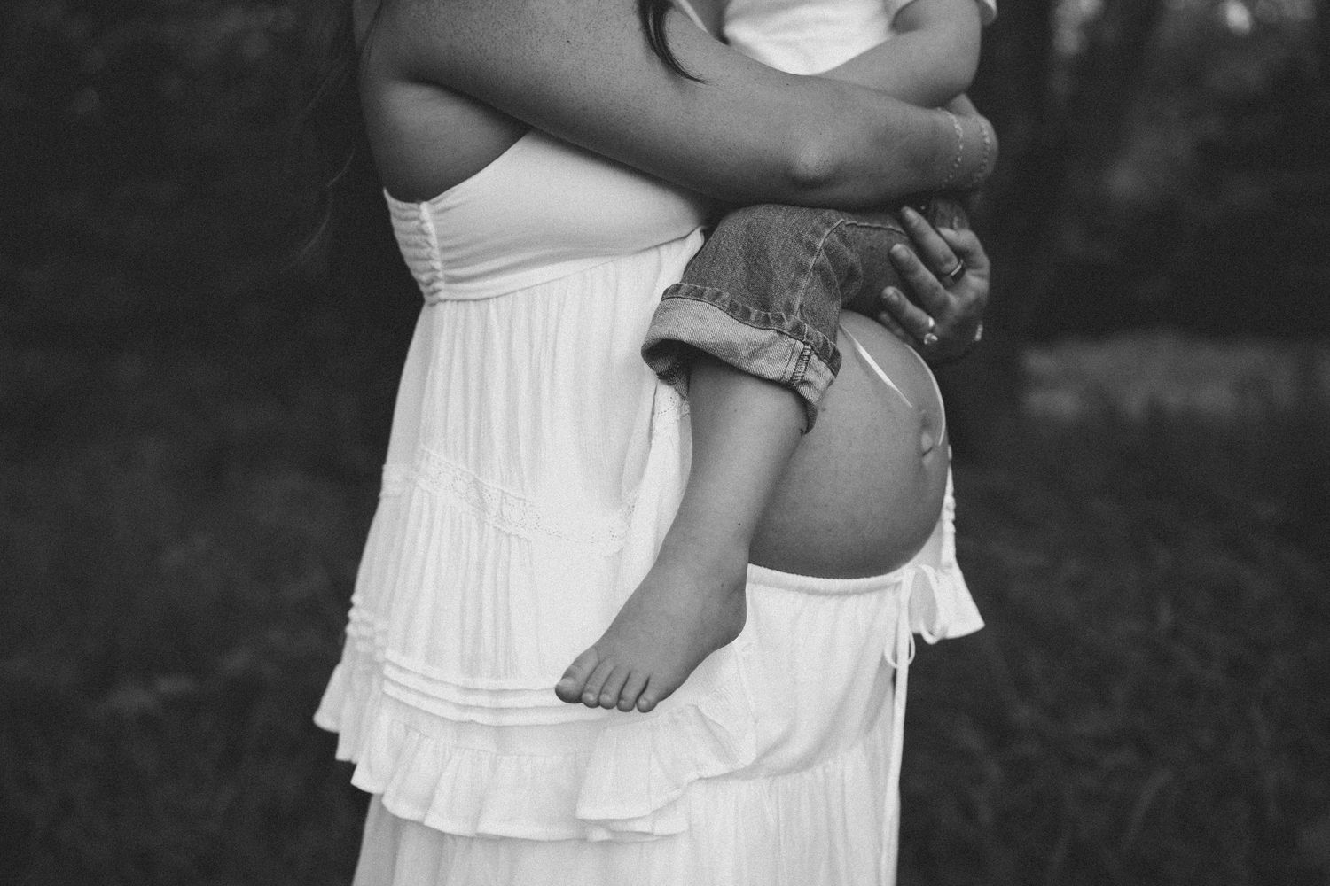 Black and white photo of pregnant woman in white dress holding a toddler outdoors.