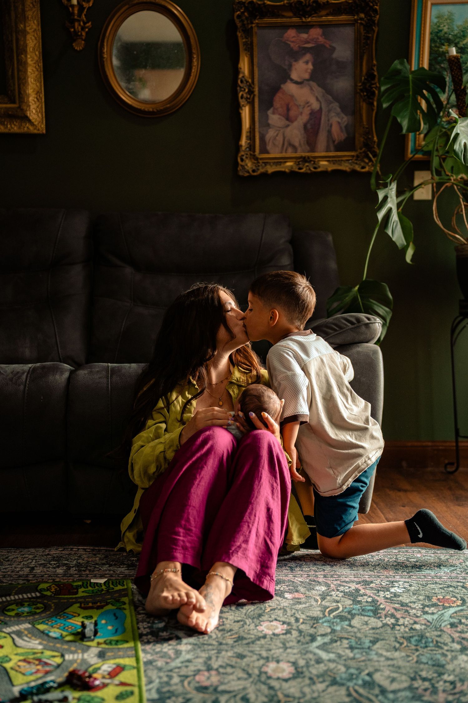 A young boy kisses his mother on the cheek as she sits on a floral rug holding a newborn baby indoors.