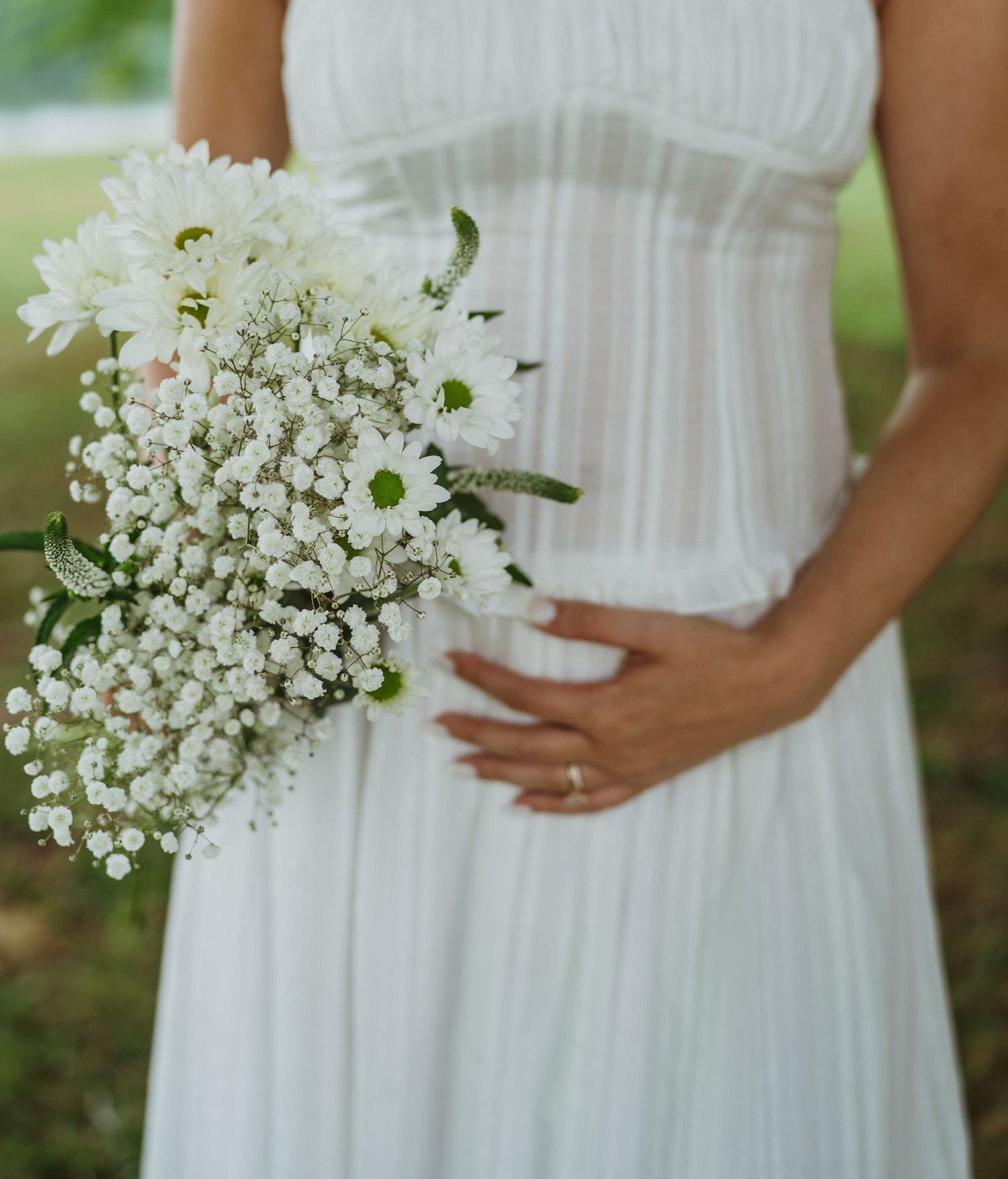 Pregnant bride in white dress holding bouquet of baby's breath and daisies outdoors.
