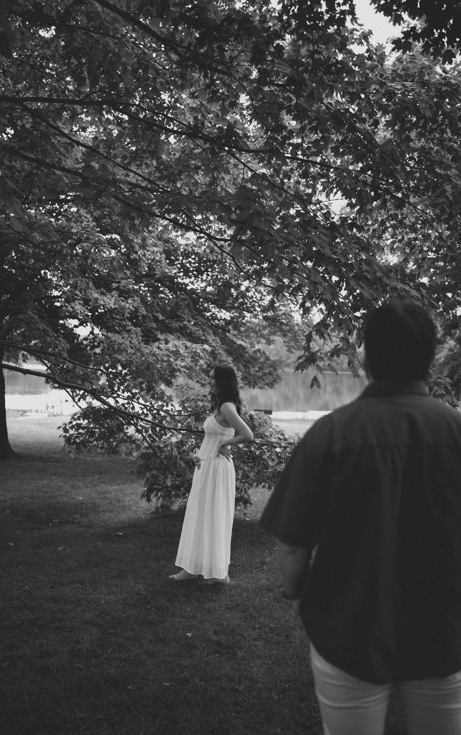 Black and white photo of couple under lush tree canopy near lake during outdoor maternity photoshoot.