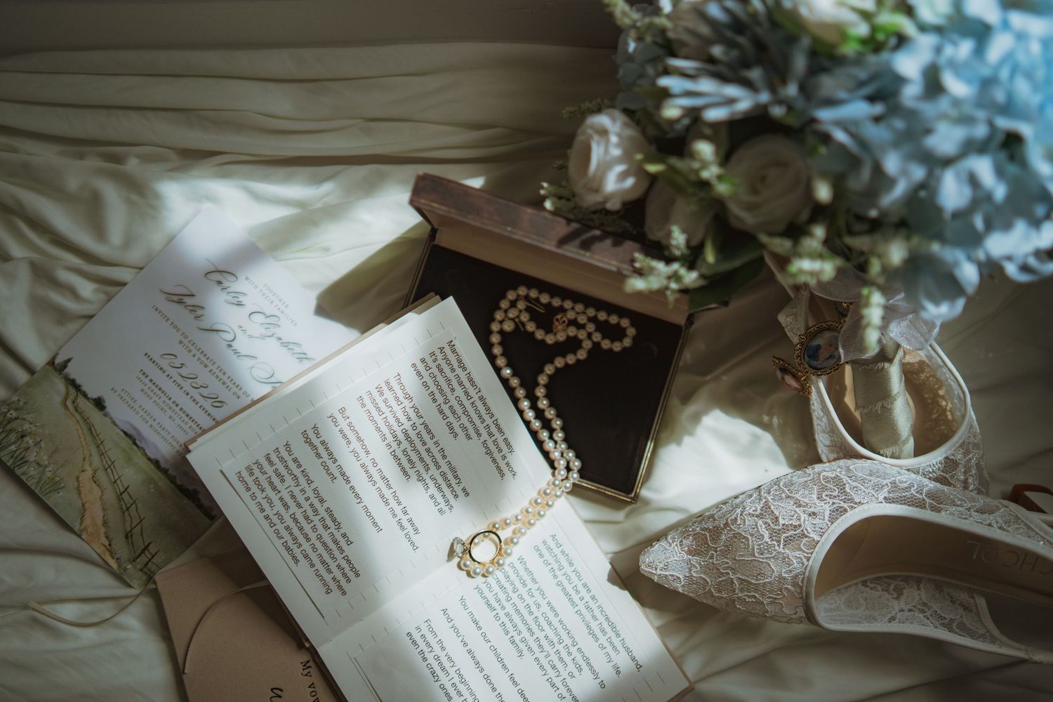 Elegant wedding flat lay with pearl necklace in box, lace heels, floral bouquet, and open book on white bedding.