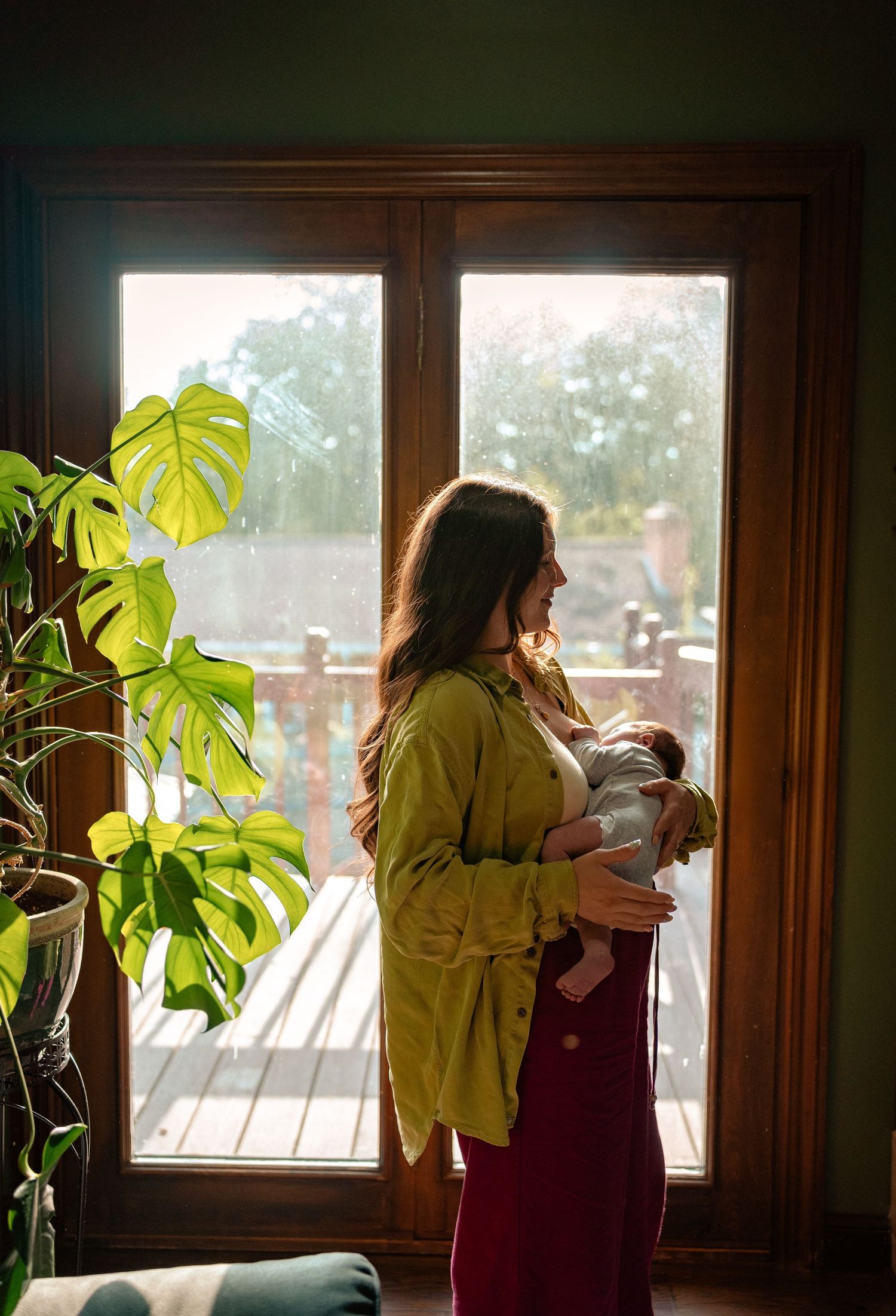 Mother in green shirt holds newborn baby by sunlit window with monstera plant.