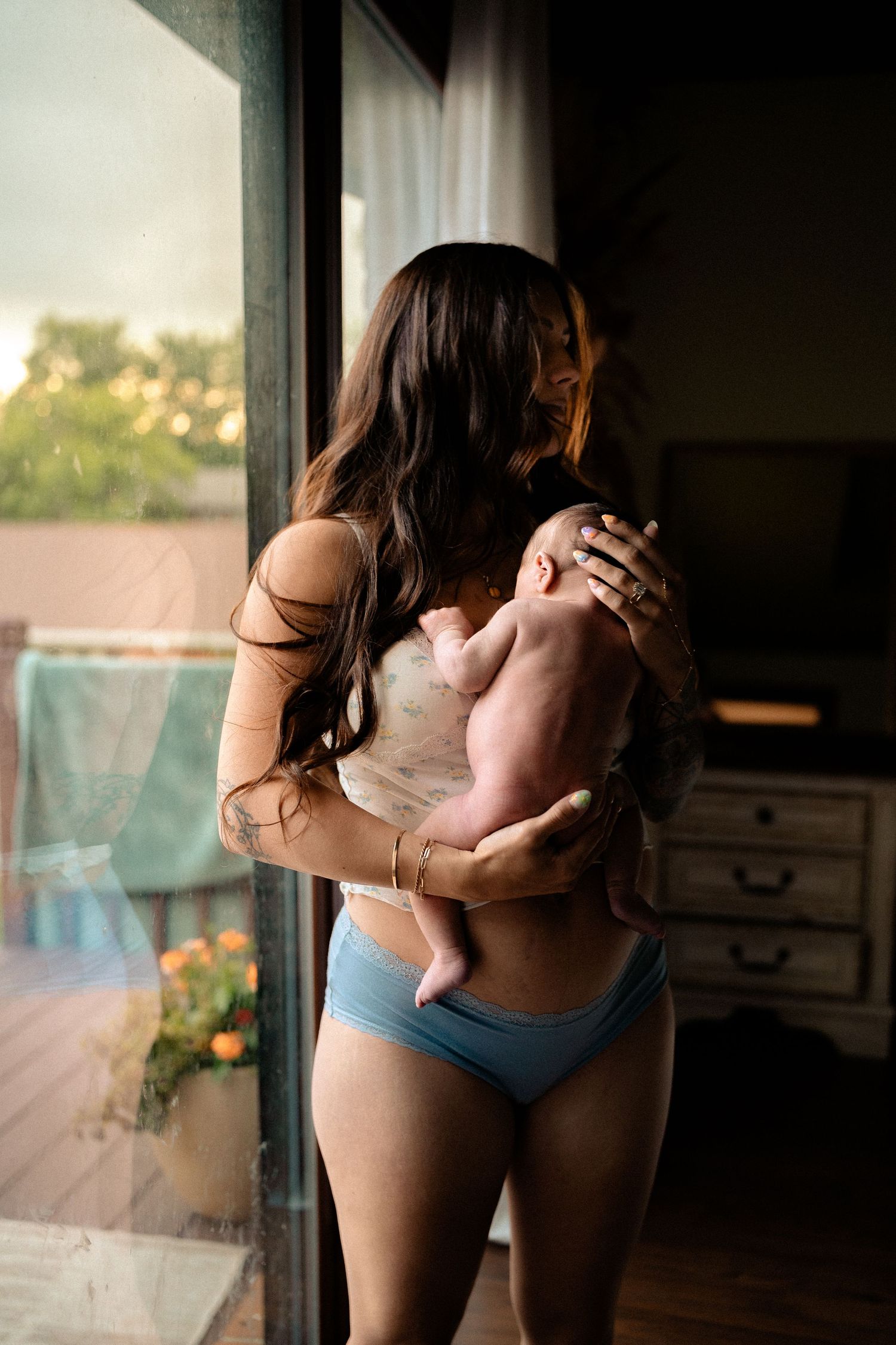 Mother in blue underwear holding newborn baby while standing by large window at golden hour.