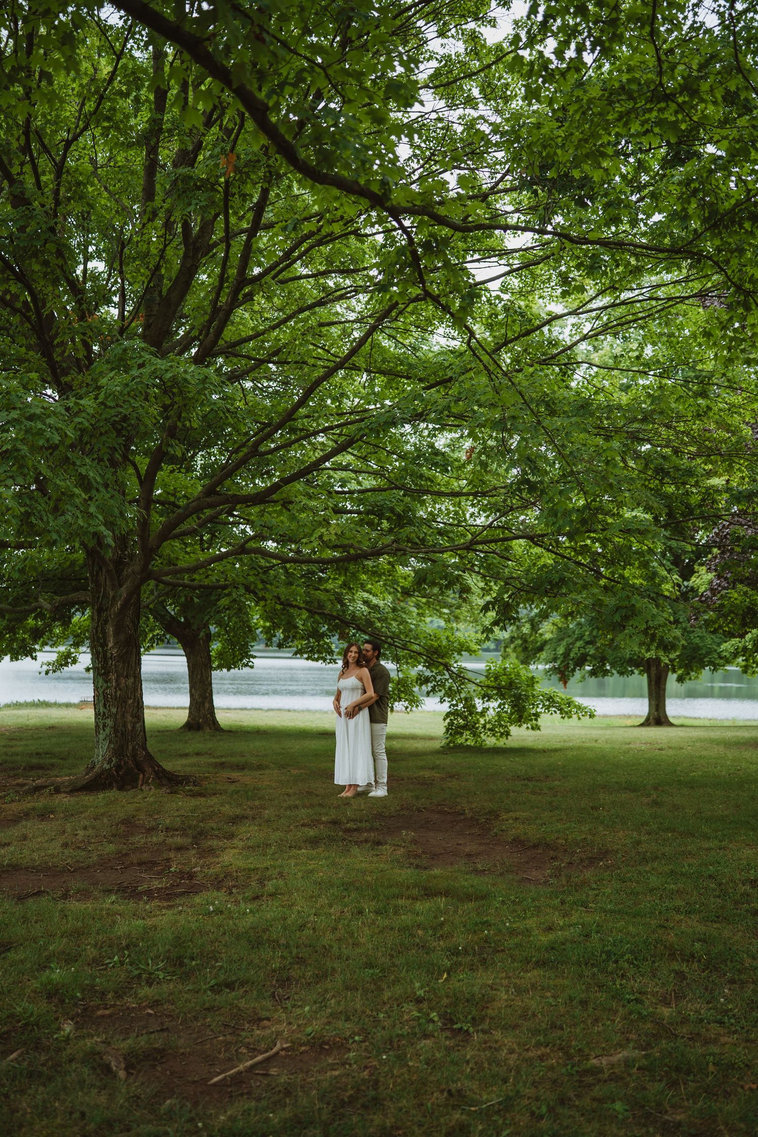 Couple embracing under large green trees in a lush park near a serene lake on a summer day.