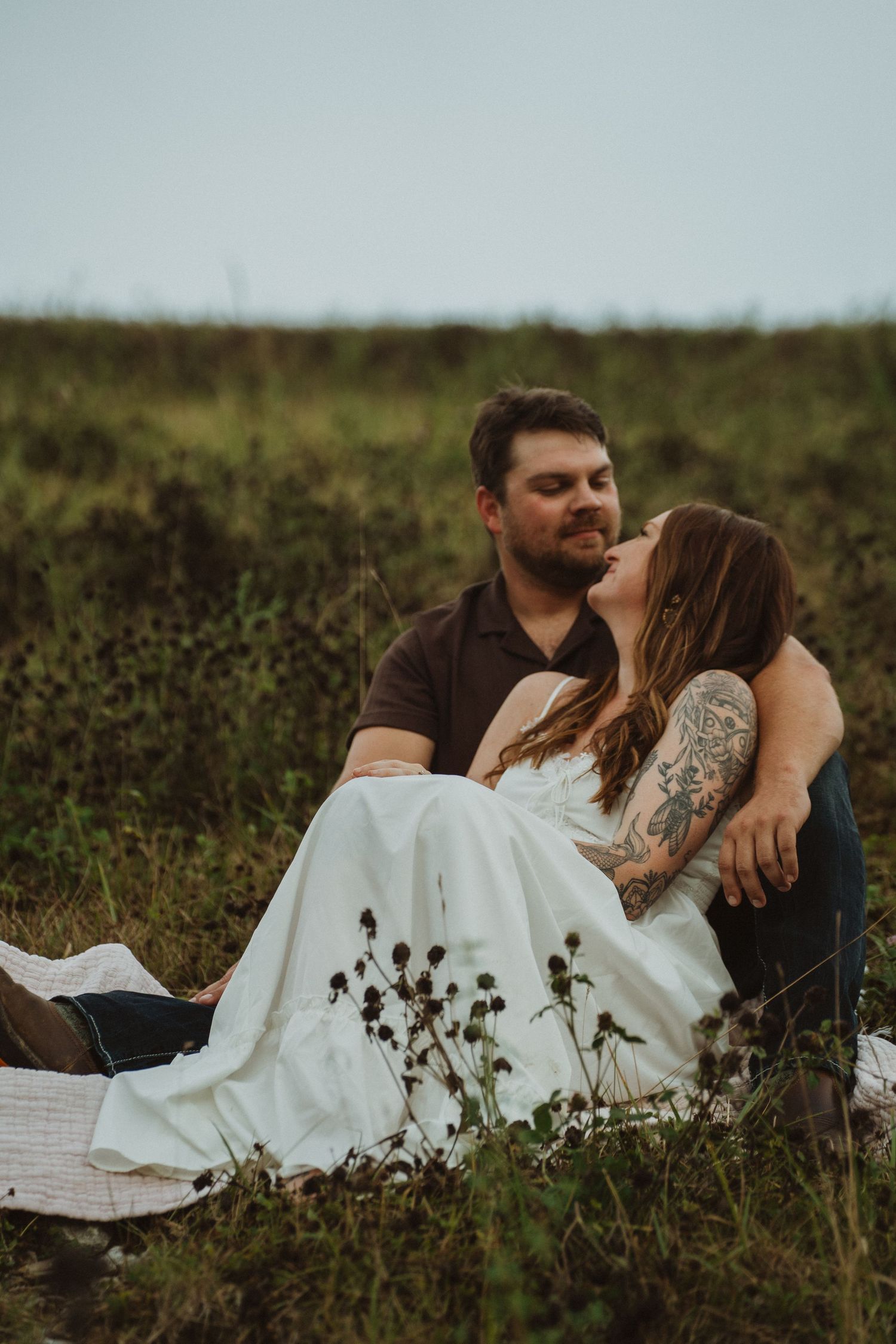 A couple shares a romantic moment sitting together in a wildflower meadow, the woman in a white dress.