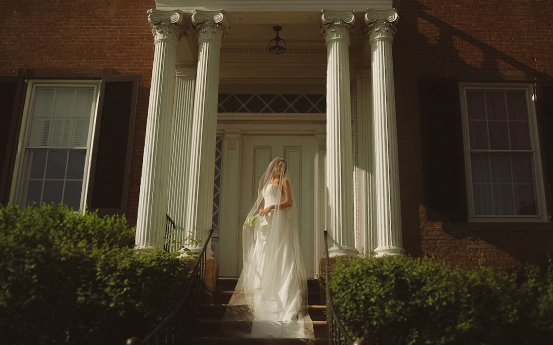 Bride in white wedding gown standing at entrance of elegant brick mansion with white columns on sunny day.