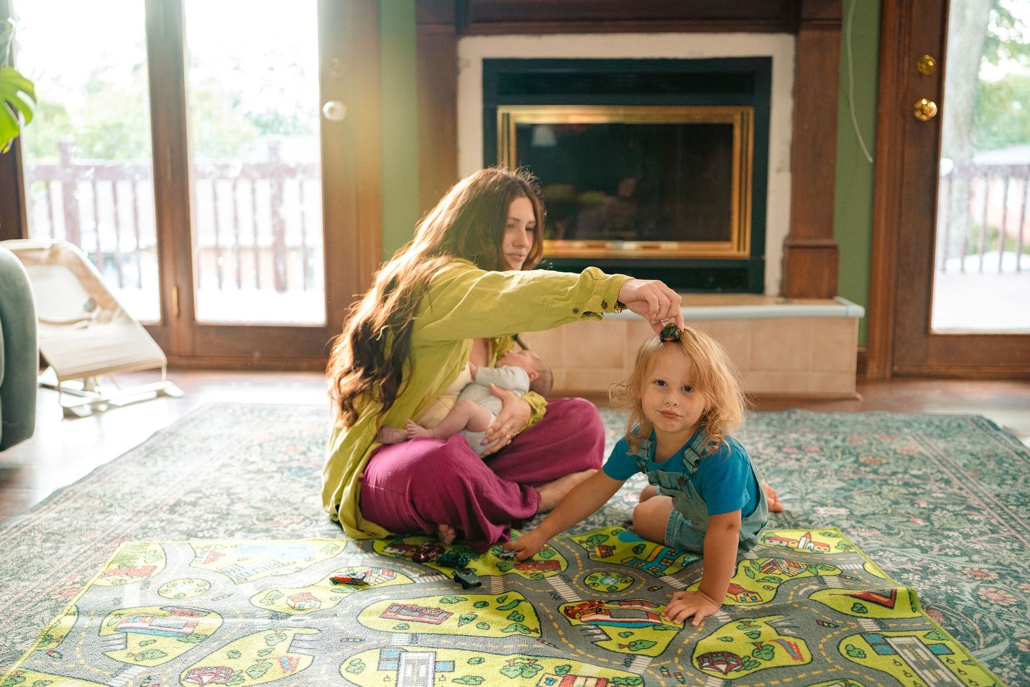 Mother playing board game on colorful rug with two toddlers by fireplace in cozy living room.