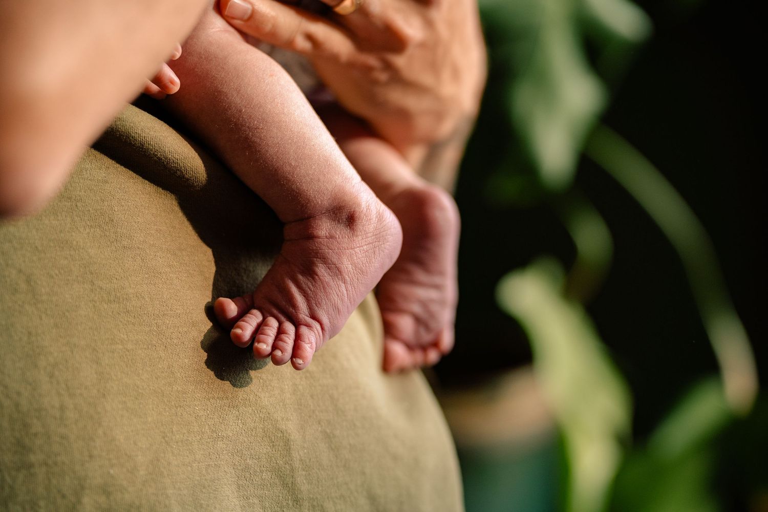 Delicate newborn baby feet and legs resting against mothers olive green clothing outdoors.