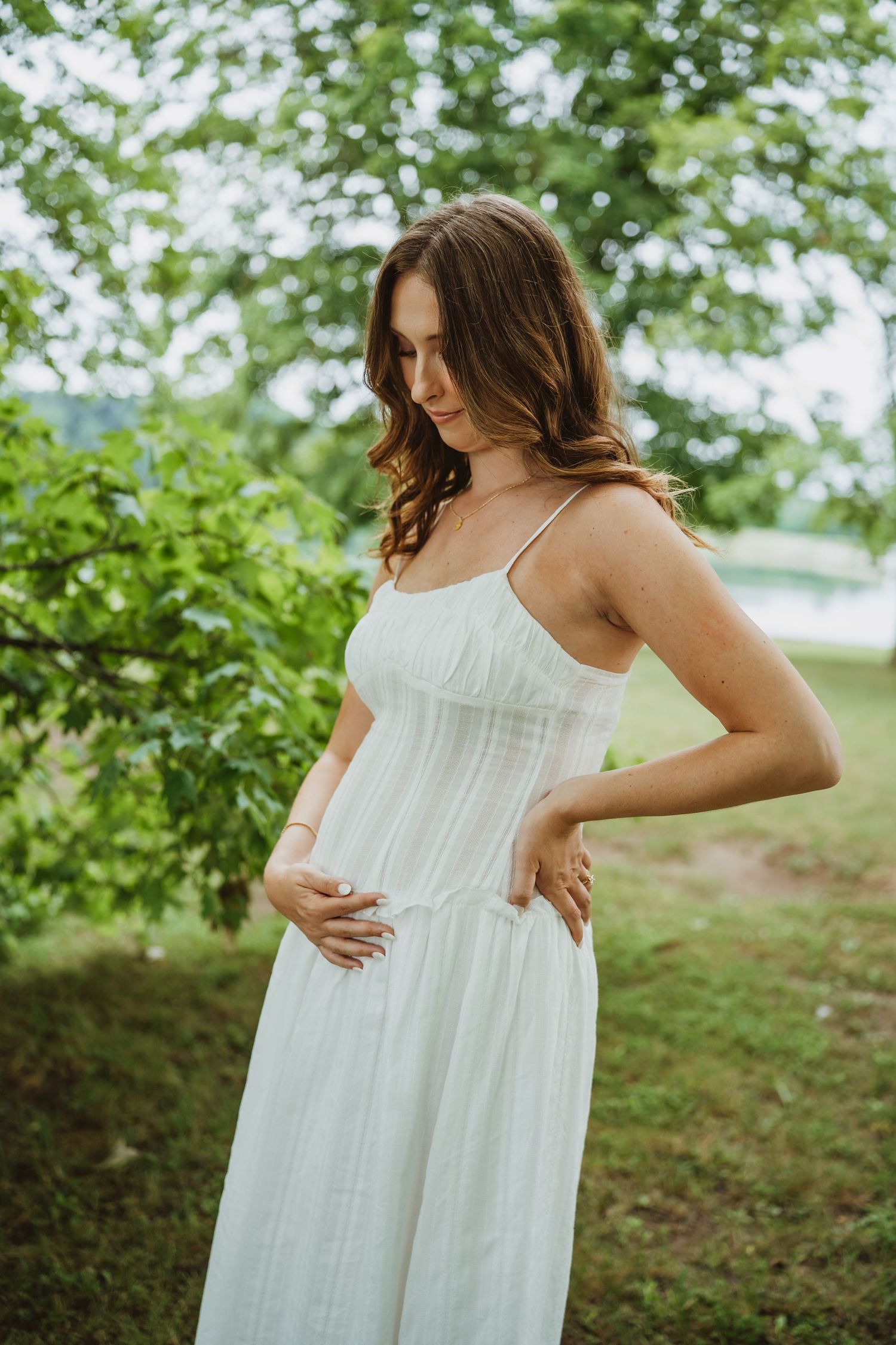 A young woman in a white sundress stands outdoors among green trees, gently cradling her baby bump.