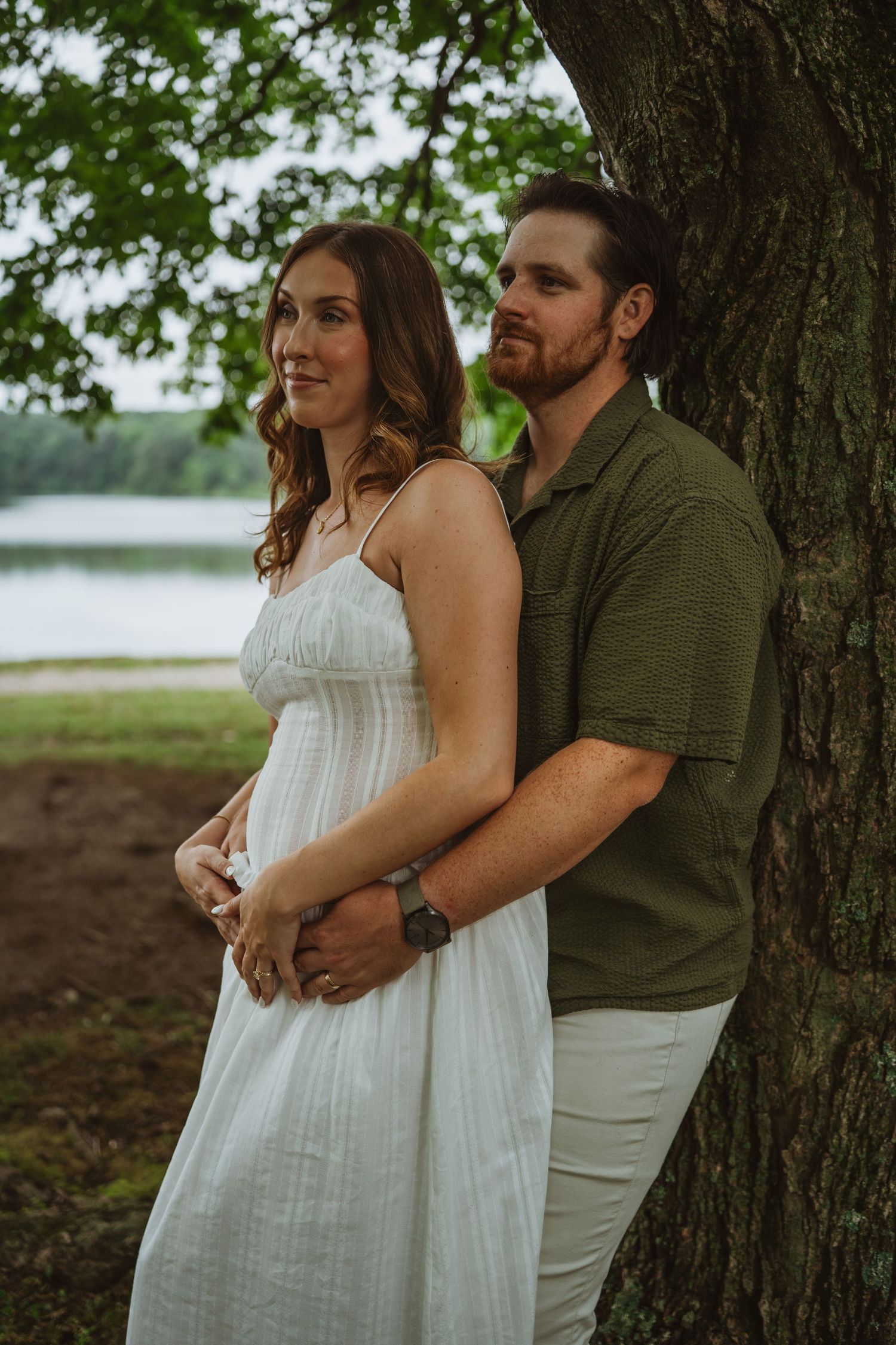 Couple embracing near a tree by a lake, woman in white dress, man in green shirt and khakis.