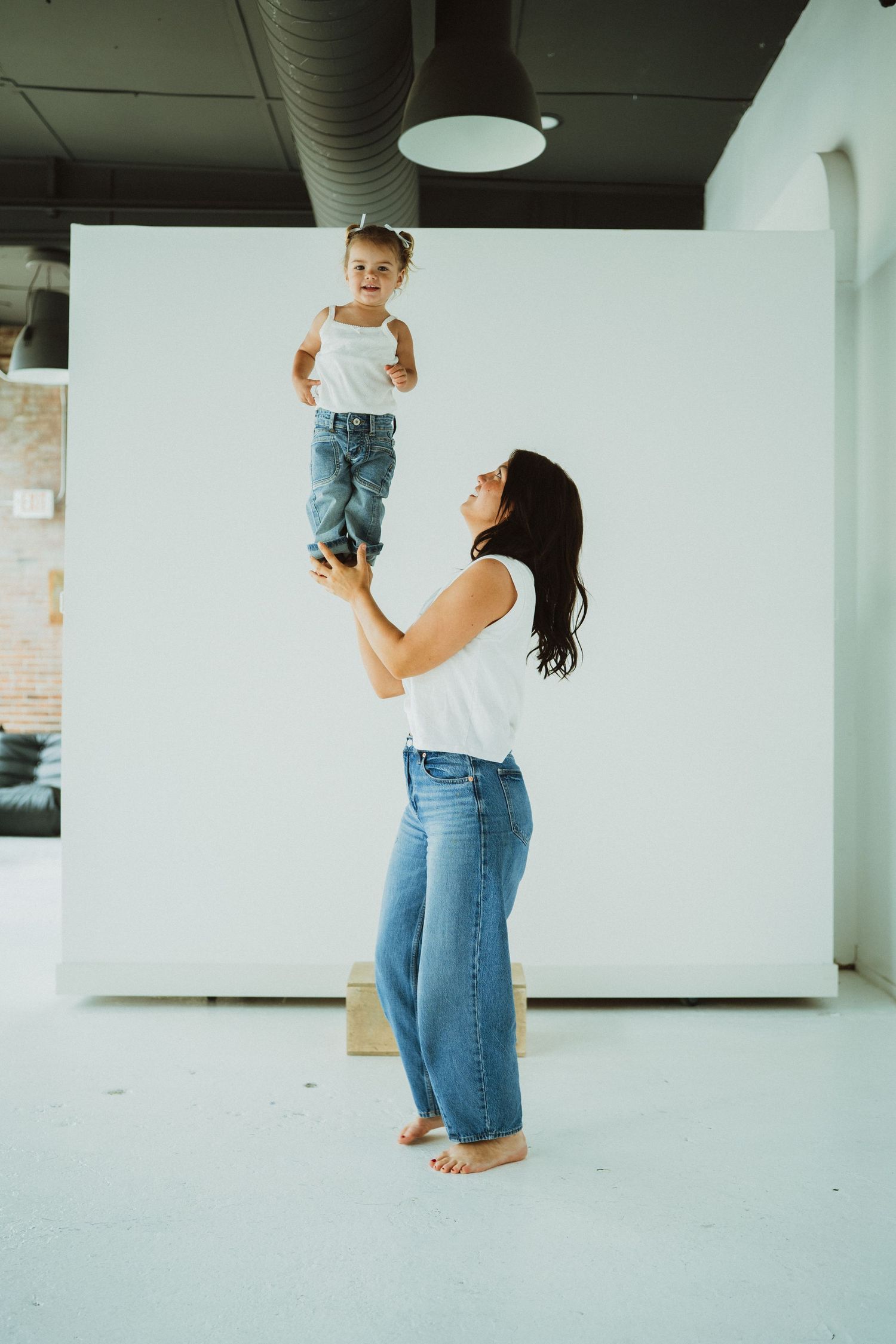 A mother joyfully lifts her toddler high in the air in a bright white studio, both wearing matching white tops and jeans.