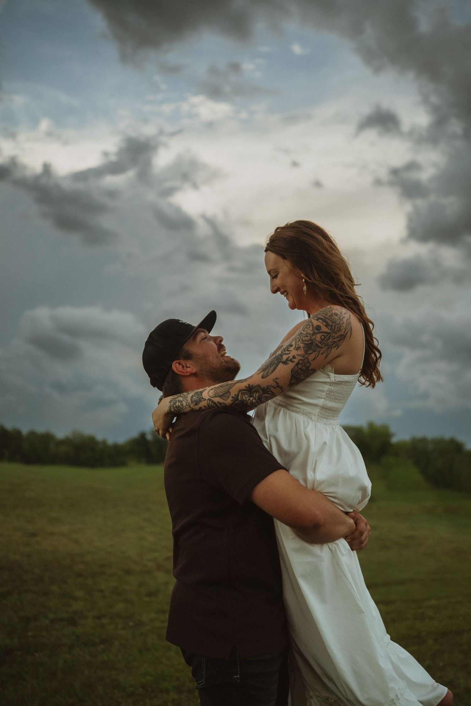 Man lifting tattooed woman in white dress under dramatic cloudy sky in an open green field.