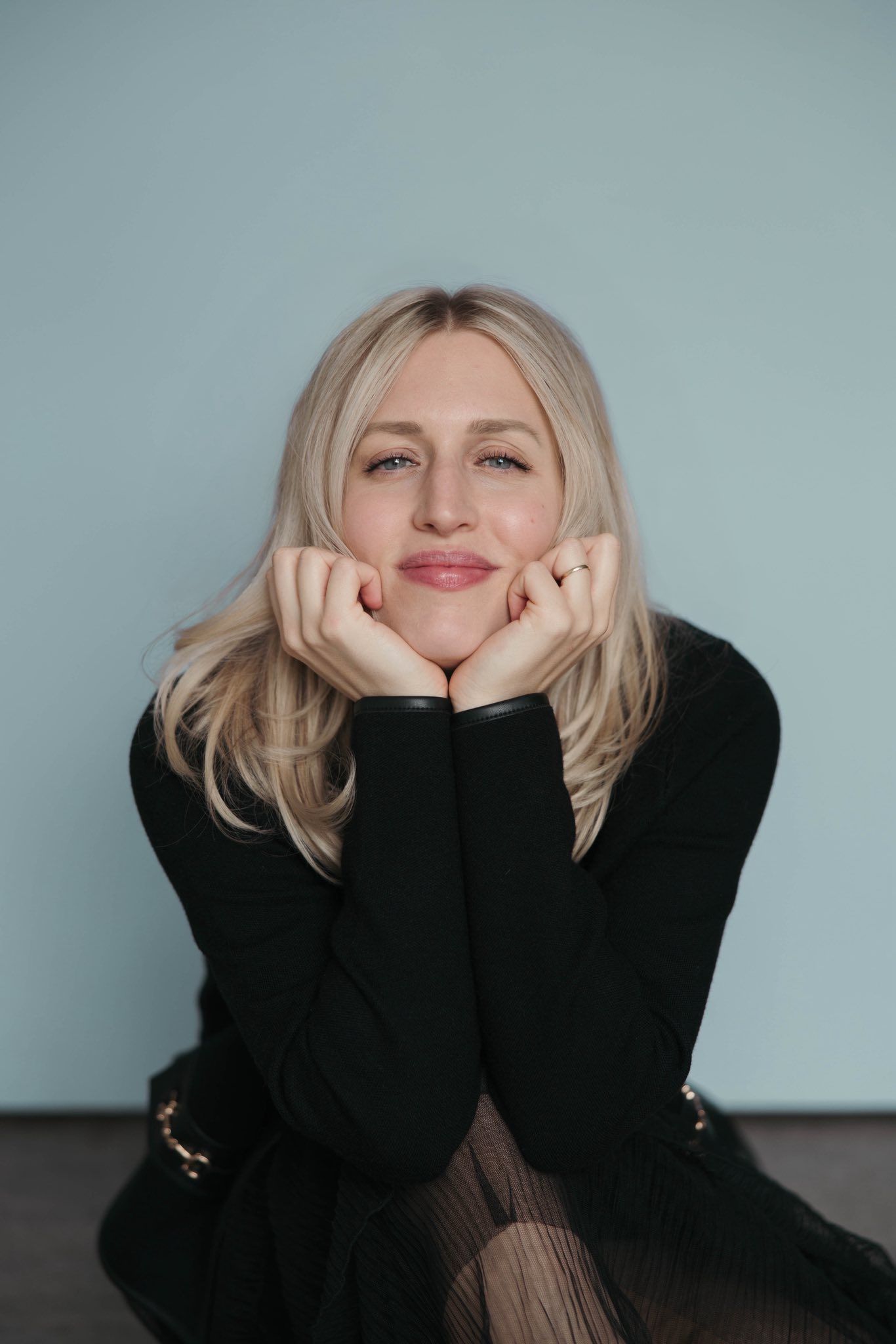 Portrait against blue wall showing blonde hair and black sweater with hands resting under chin in casual pose.