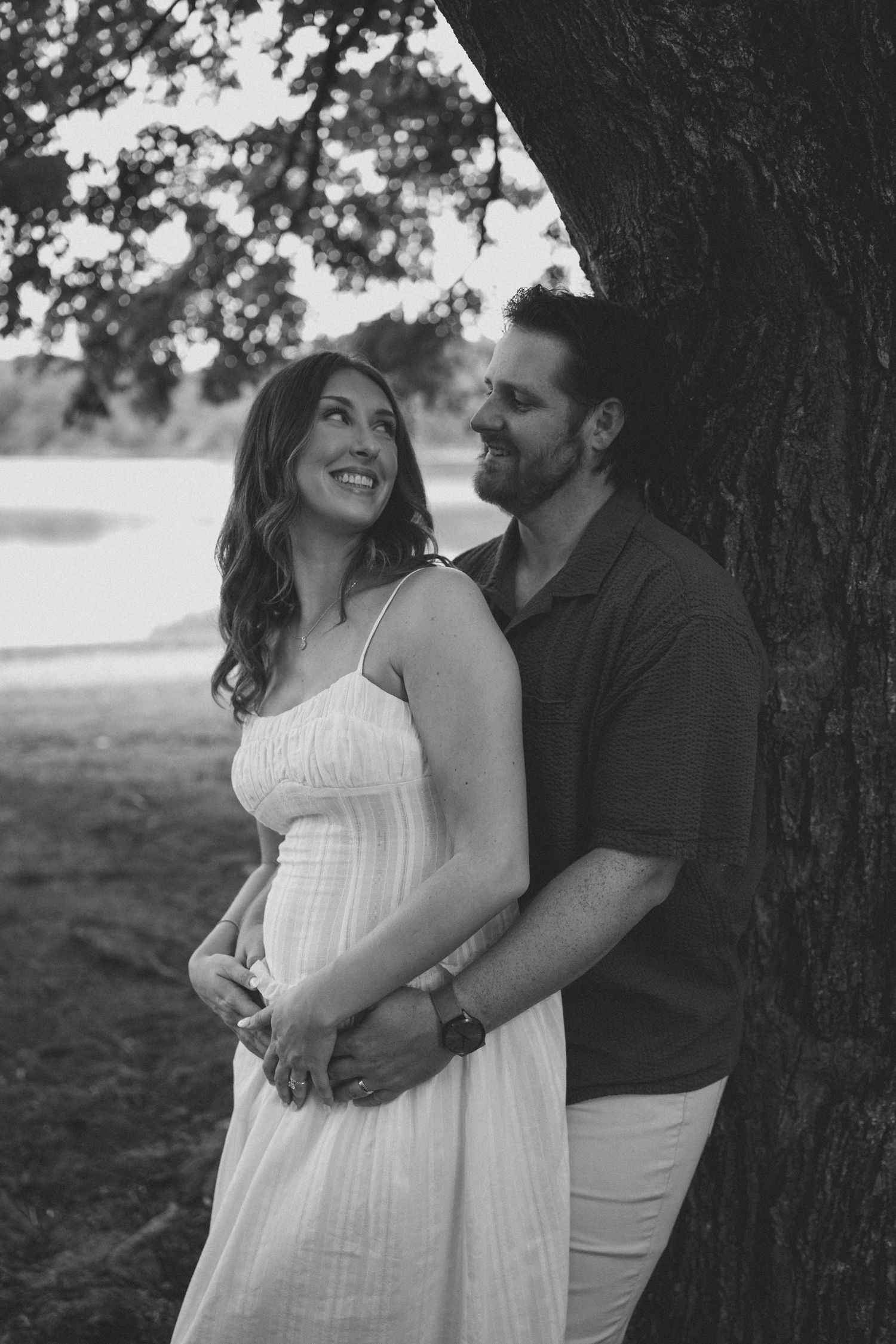 Black and white photo of pregnant woman in white dress embraced by man leaning against tree by lake.