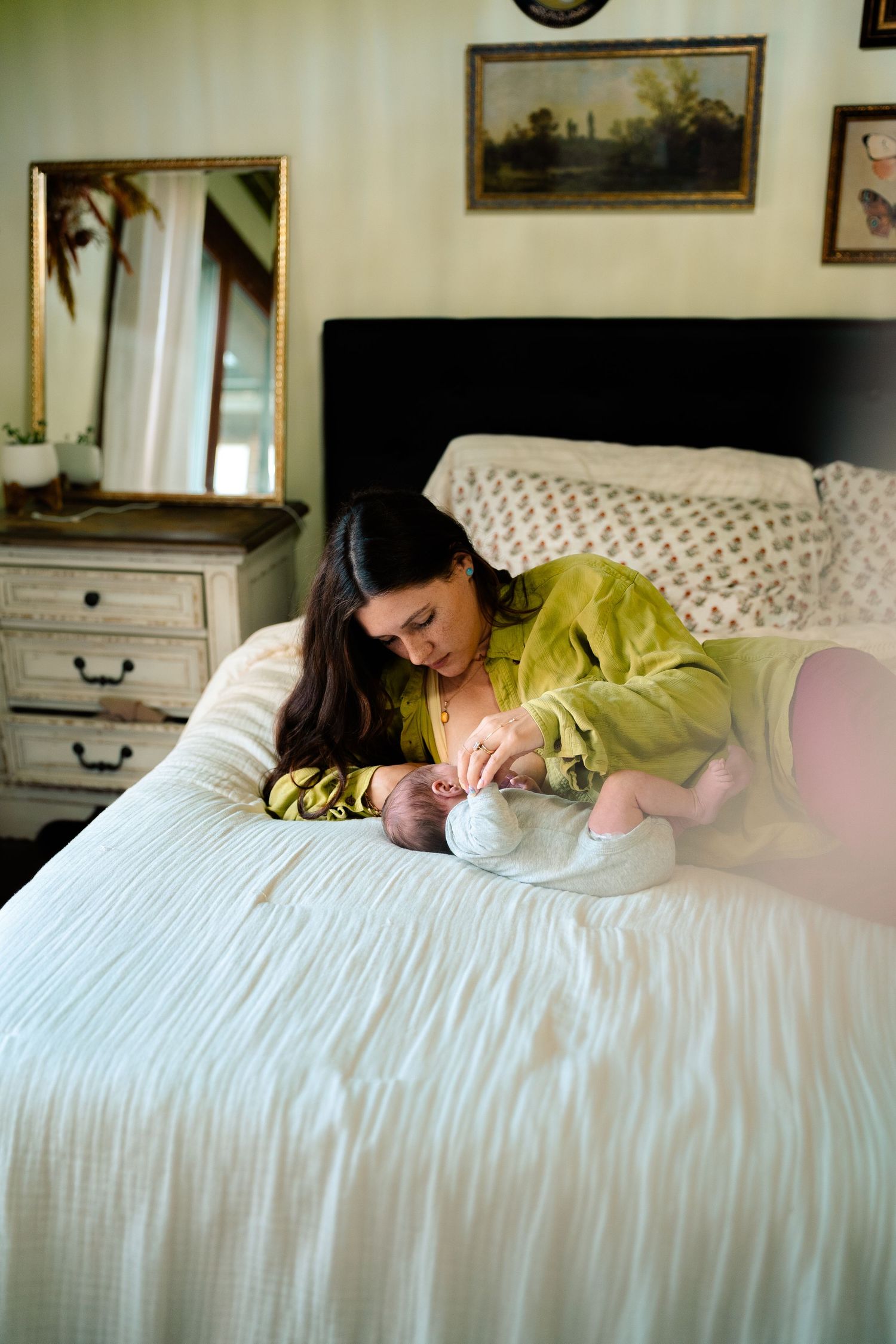 Mother in green top leans over newborn baby on white bed in cozy bedroom.
